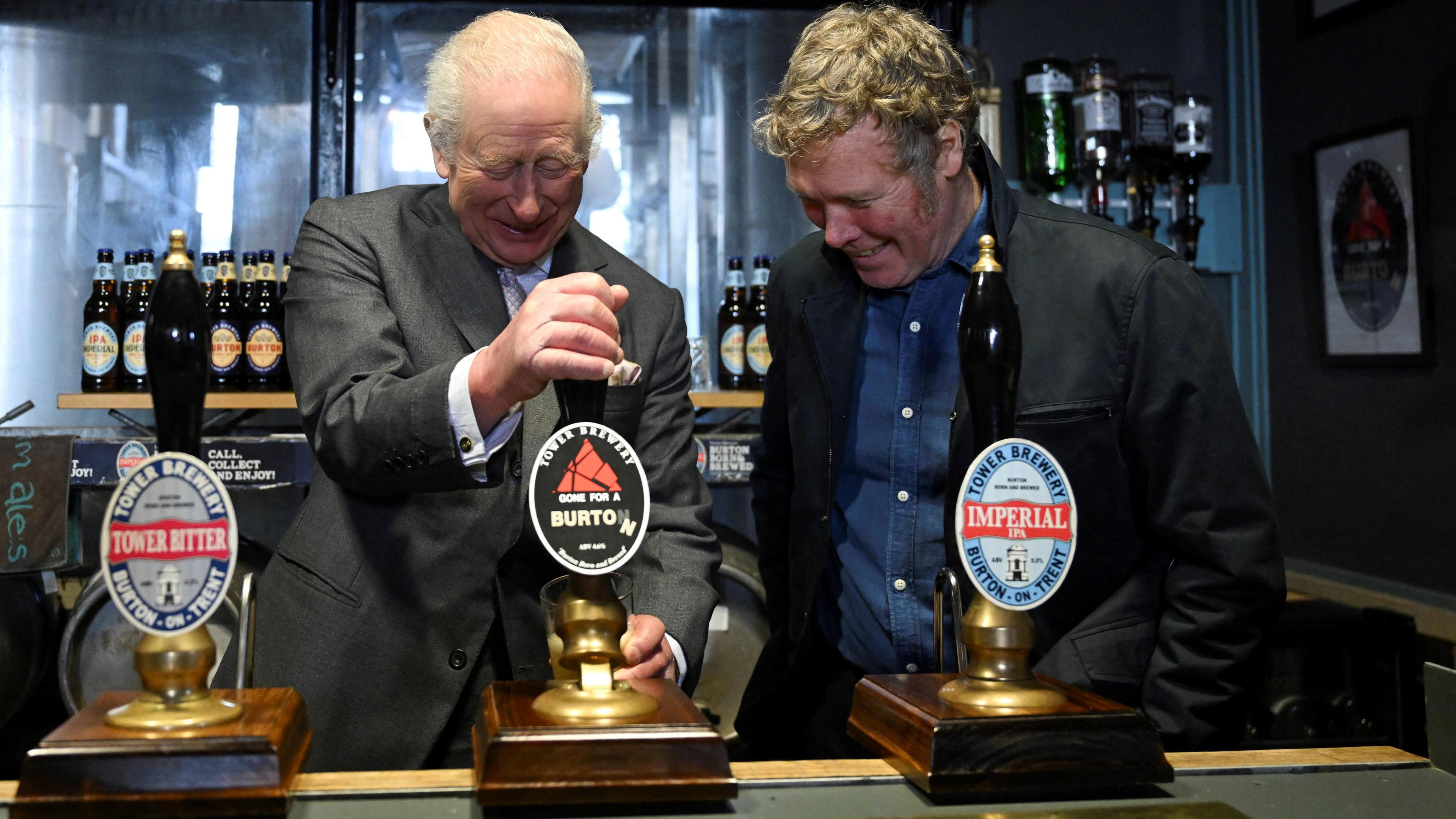 King Charles, wearing a grey suit, stands behind a bar and pulls a pint while a man wearing a black jacket and blue shirt stands next to him and watches.