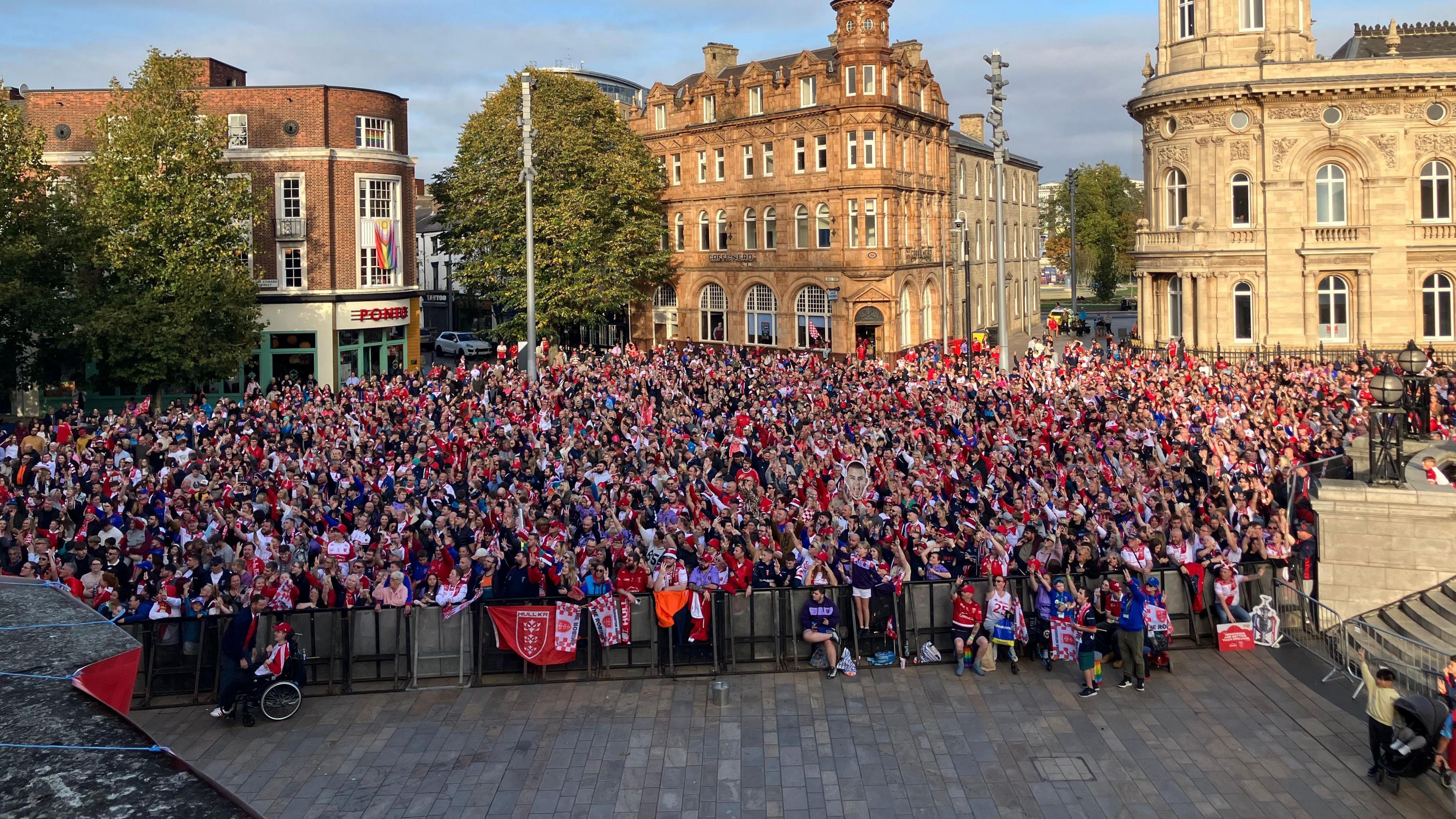 Thousands of fans in red tops, hats and scarves are stood behind a barrier.