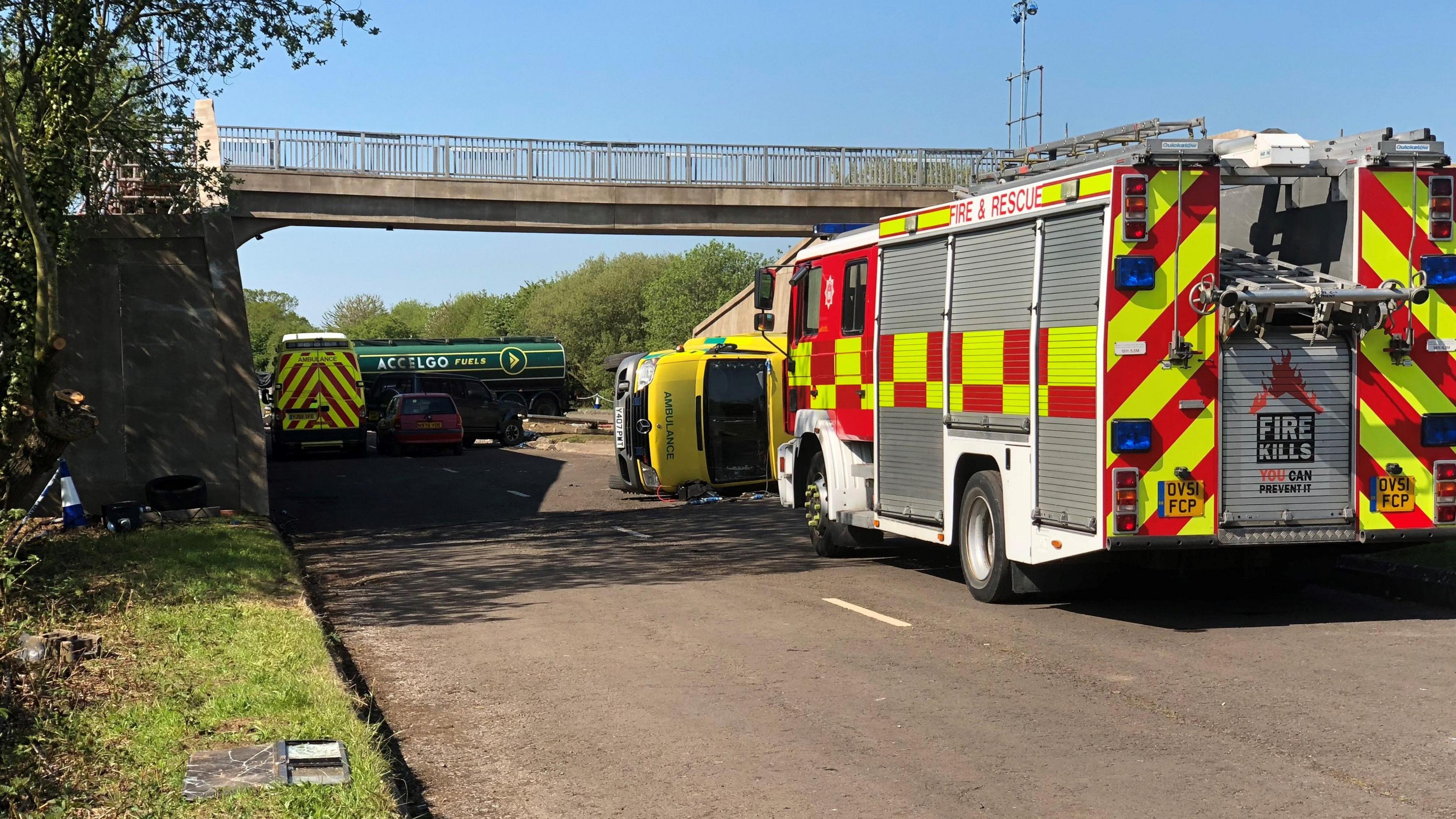 A fire and rescue truck is pictured on an empty dual carriageway. In front of it is an ambulance on its side. In the distance is a fuel lorry.