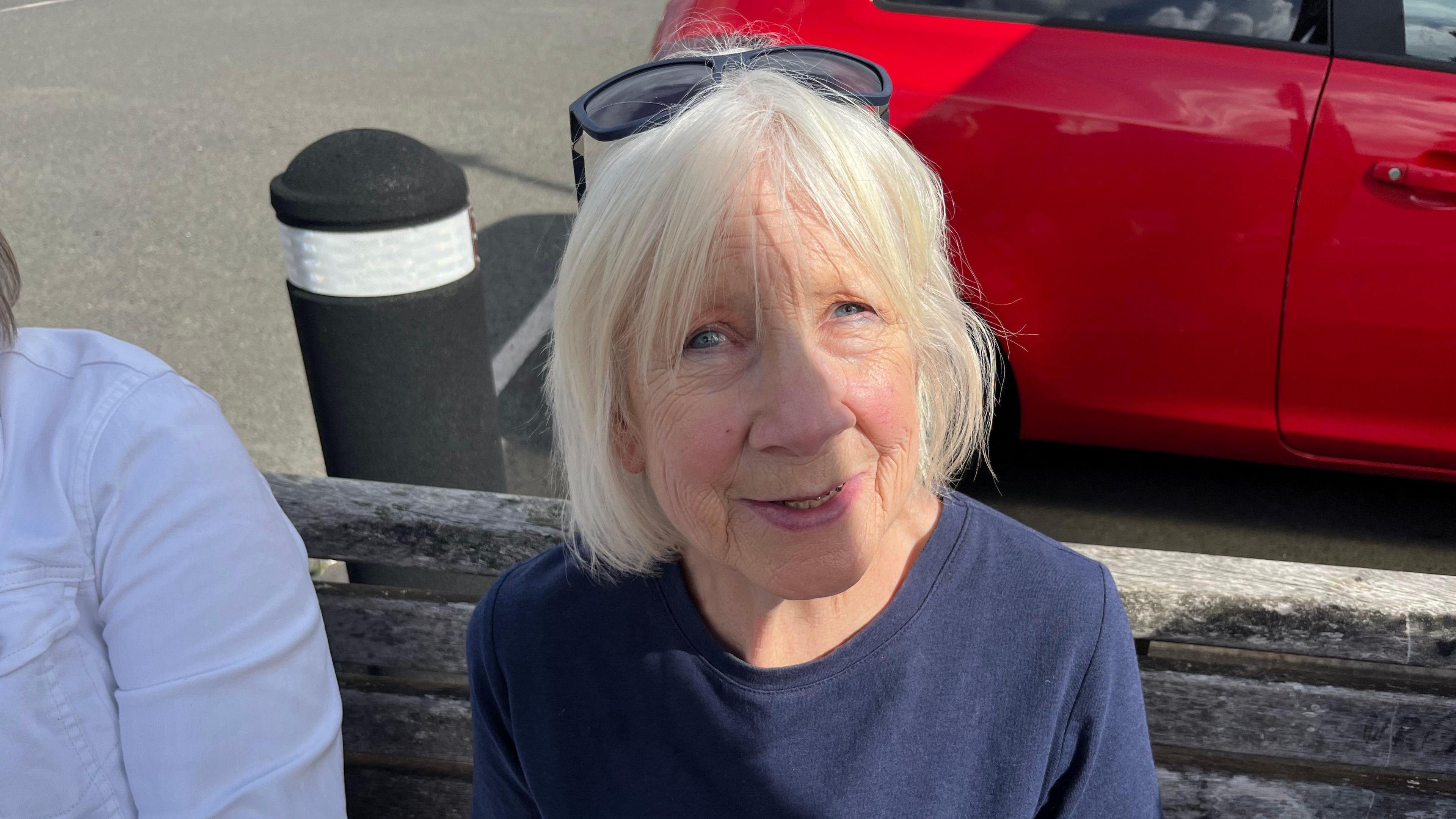 Ms Warren is sitting on a wooden bench as she waits for the bus. She has medium length white hair and blue eyes. She is wearing sunglasses on top of her head and has on a navy jumper.