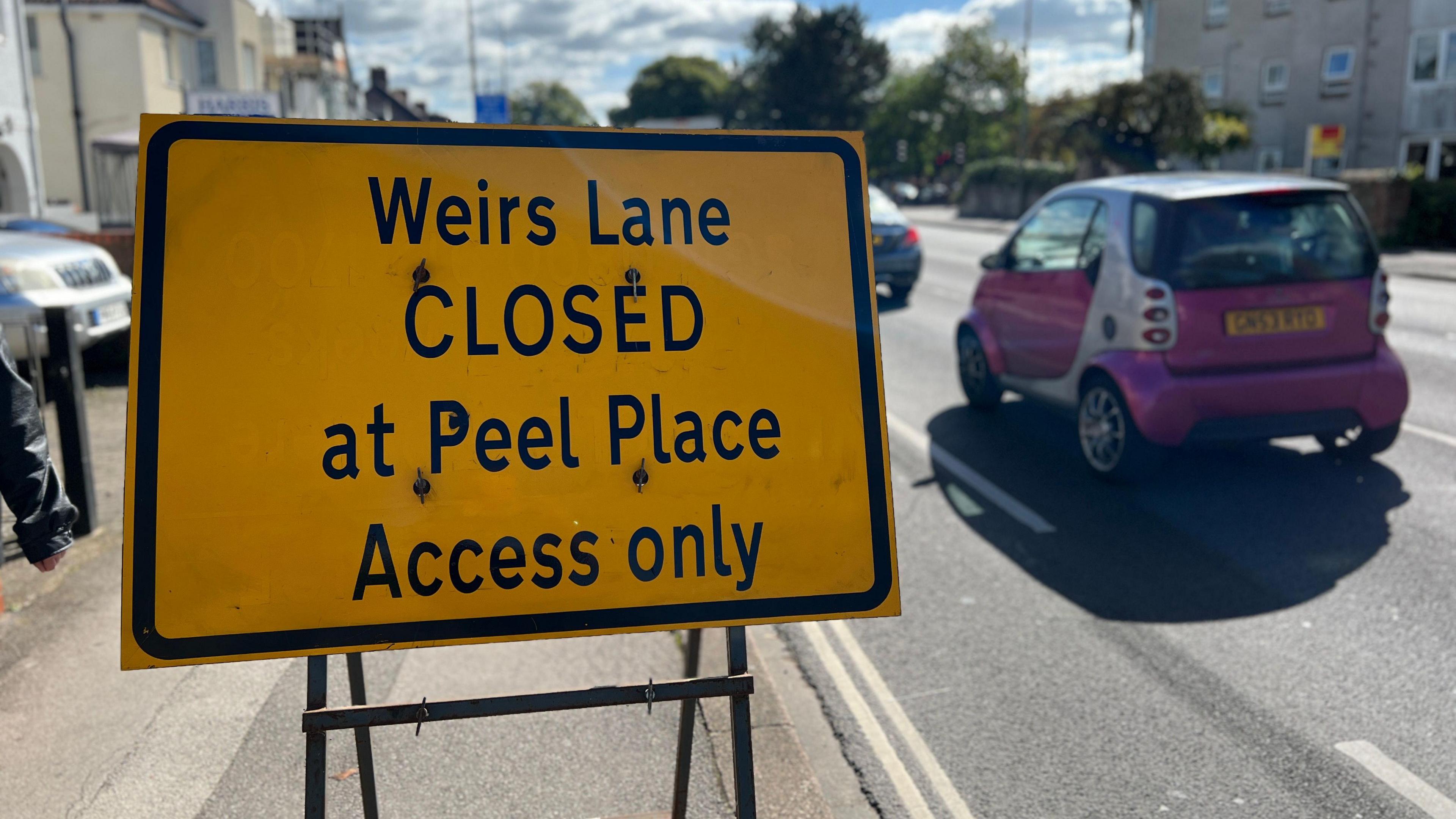 A  close up of a yellow street sign reading Weirs Lane CLOSED at Peel Place Access only. A car could be seen on the road behind it. It is a sunny day.