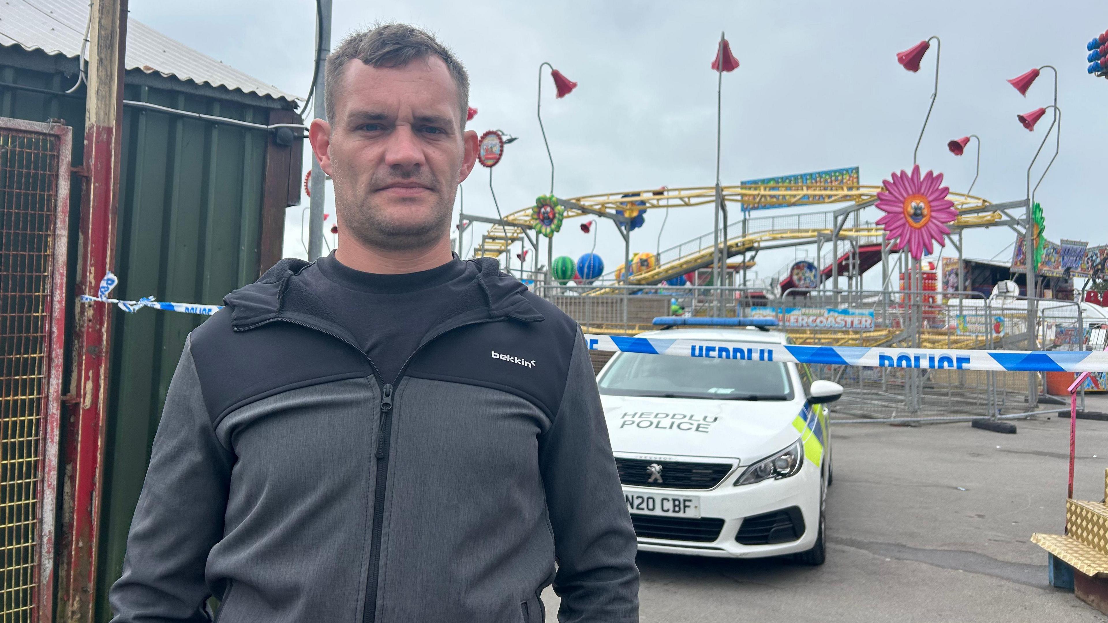 John, who has short brown hair and stubble, wearing a black and grey hoodie. There is a police car behind him in front of a police cordon. In the background the amusement park can be seen with a rollercoaster track fenced off by metal gates.