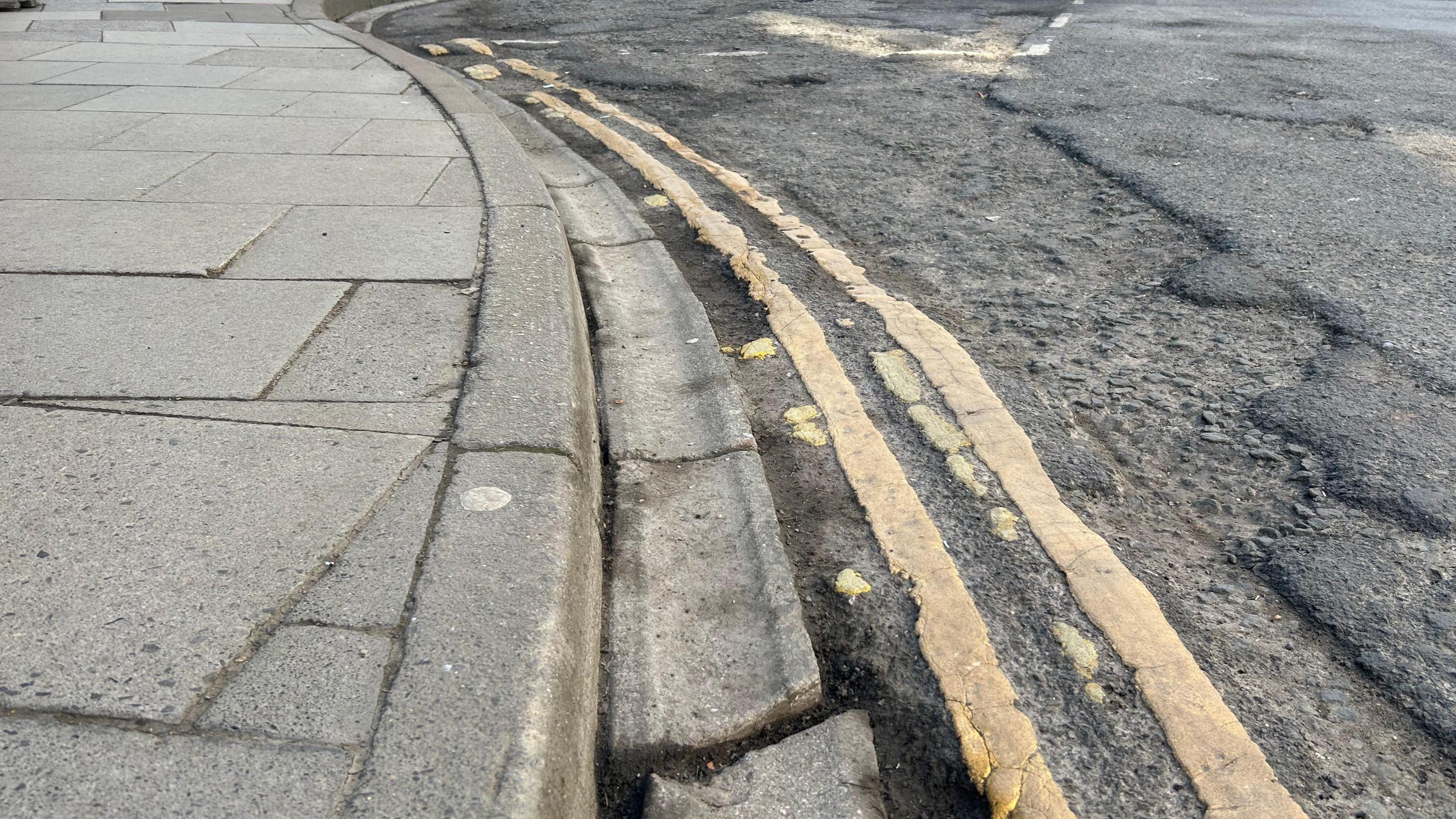 A Wiltshire road with an uneven surface with the top layer of tarmac broken up. The gutter running next to the pavement is broken and the yellow lines are patchy