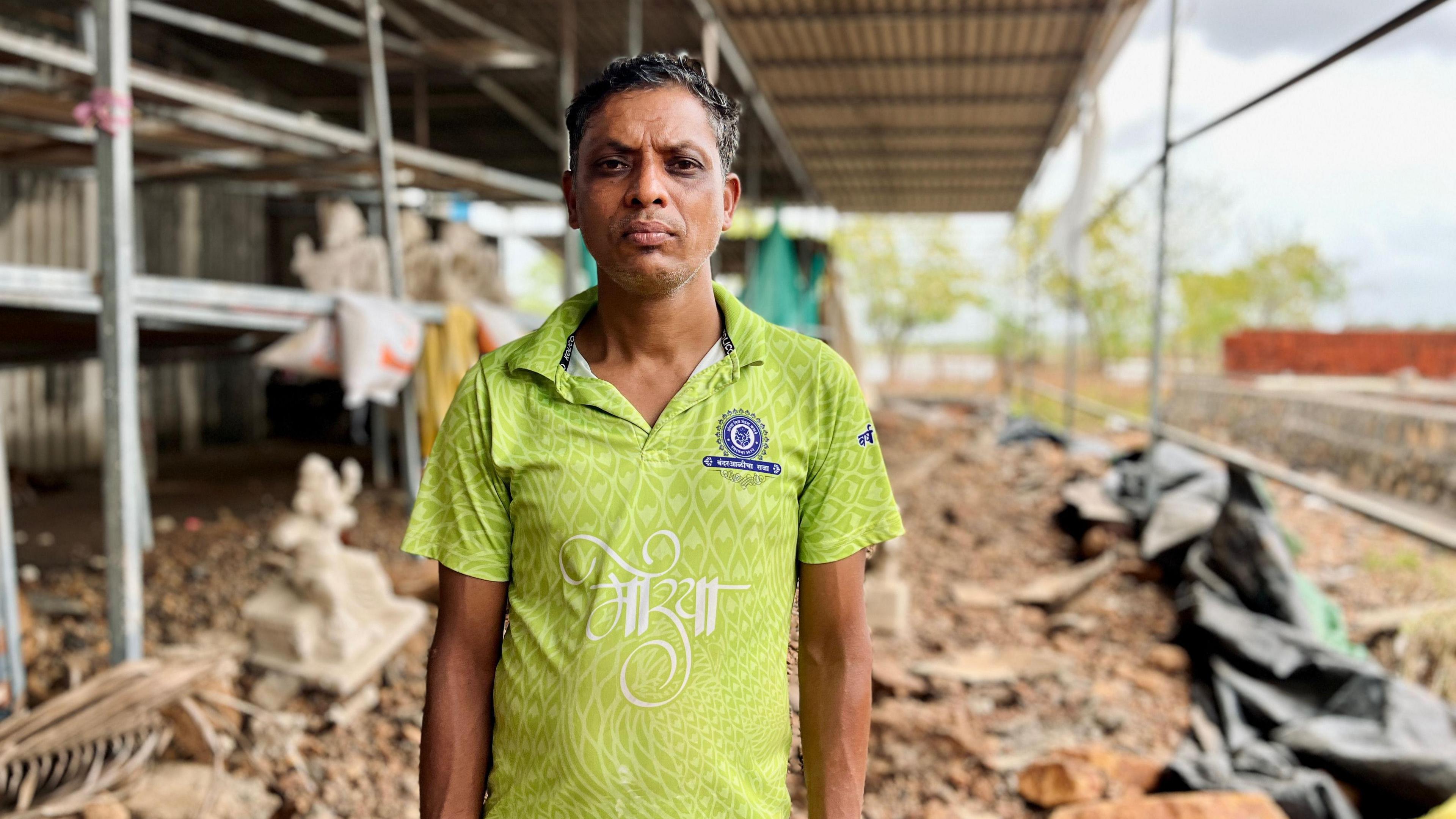 Dilip Mhatre stands in a green t-shirt, behind him is a blurred backdrop of his damaged workshop