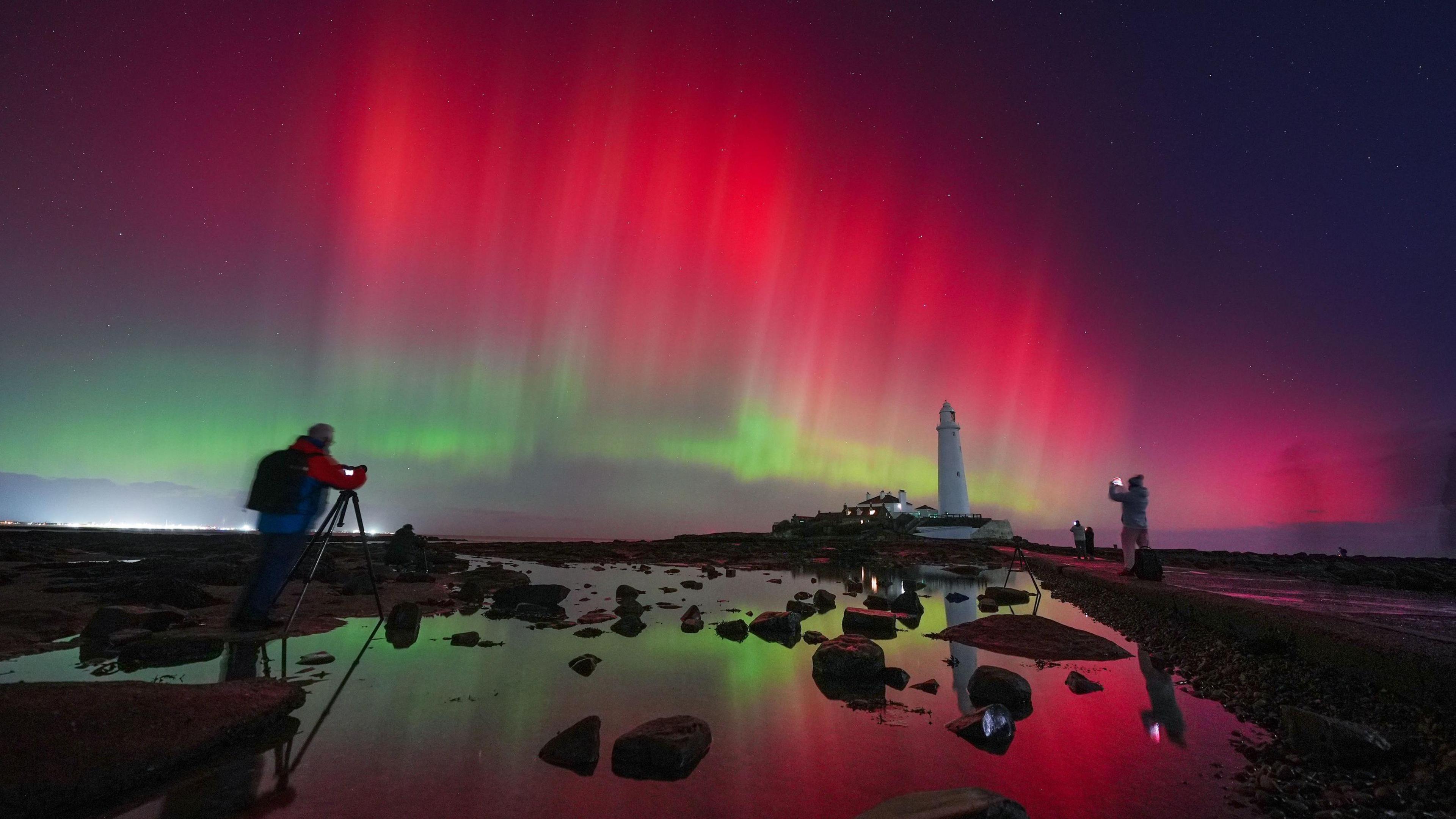 Whitley Bay lighthouse in the distance, with the bright colours of the Northern Lights—reds and greens—shining in the sky. Some of the light is reflected on the water in the foreground, and a number of people can be seen taking photographs.