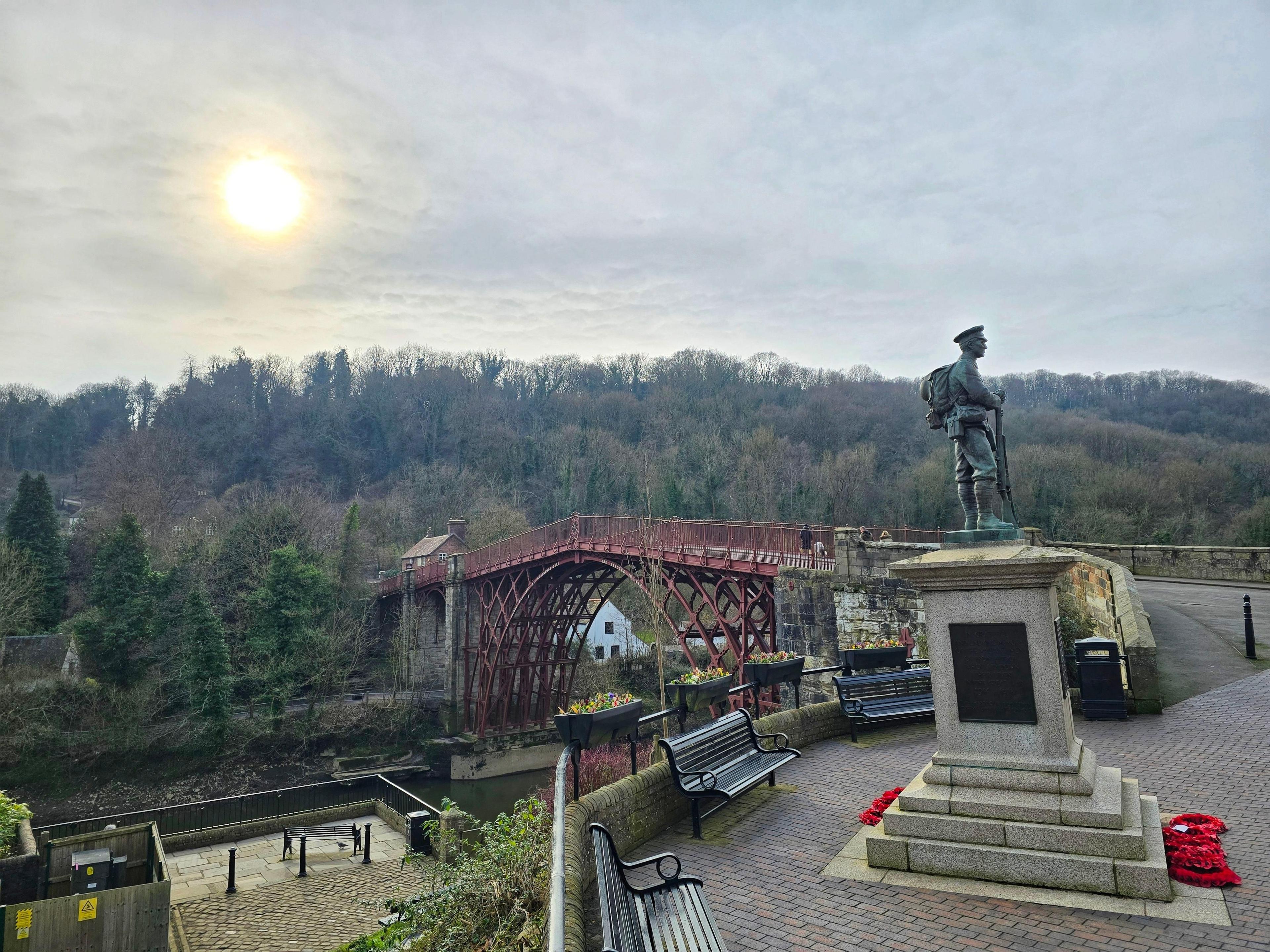 18th Century arched iron bridge painted in a deep red, stretching over a deep gorge. In the foreground, a statue of a soldier cast in metal atop a stone plinth with memorial poppy wreaths at its base.