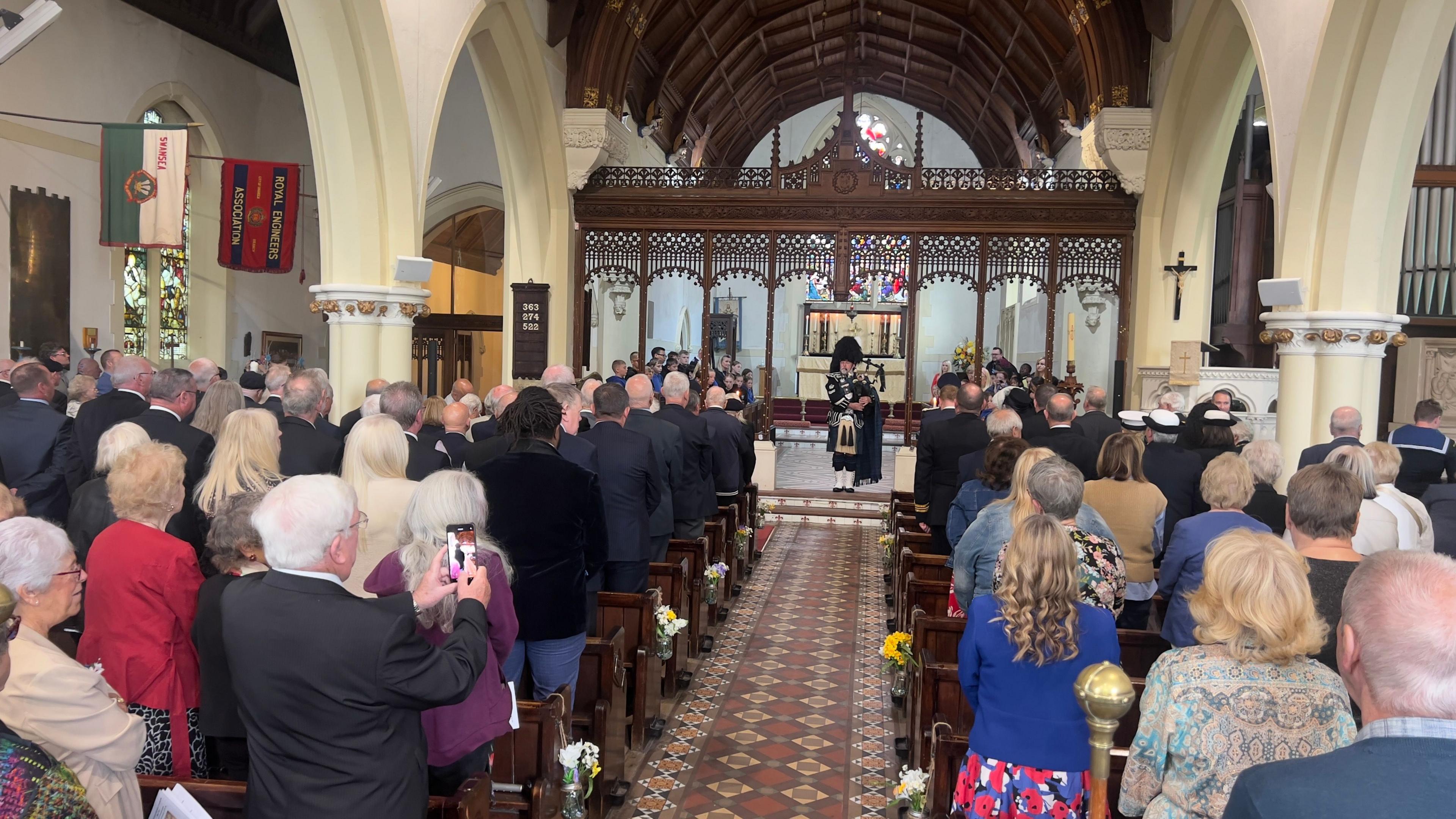 People in church pews seen from behind standing up with man playing bagpipes at the front