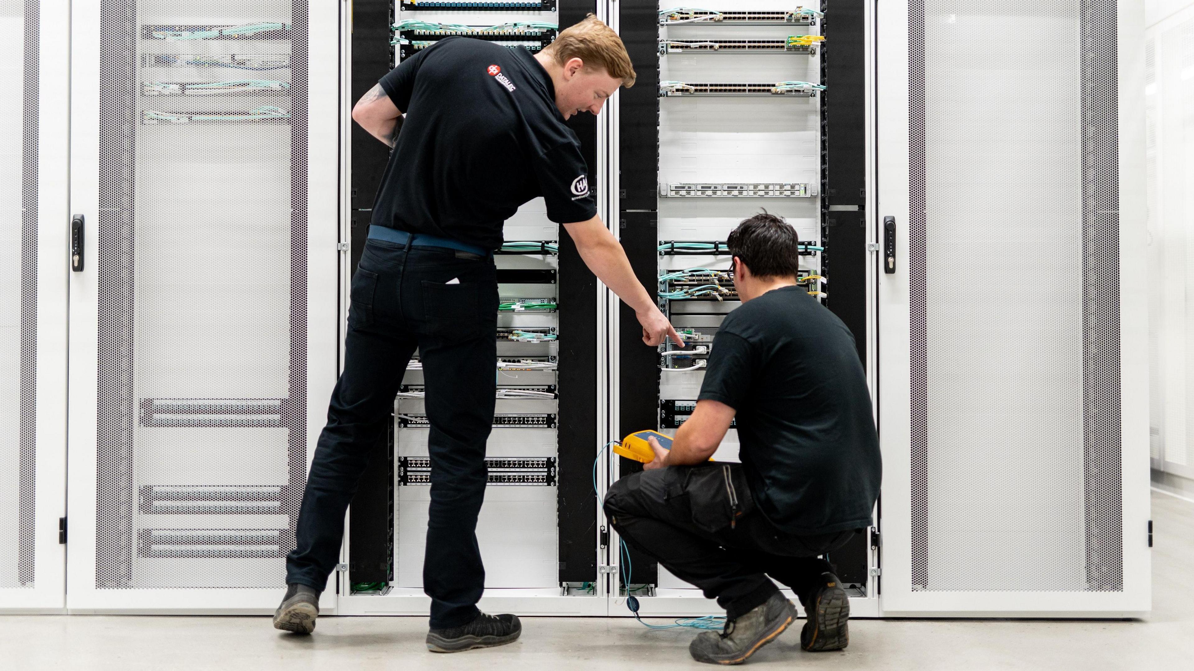 Two male engineers in black outfits work on wiring on racks in a datacentre