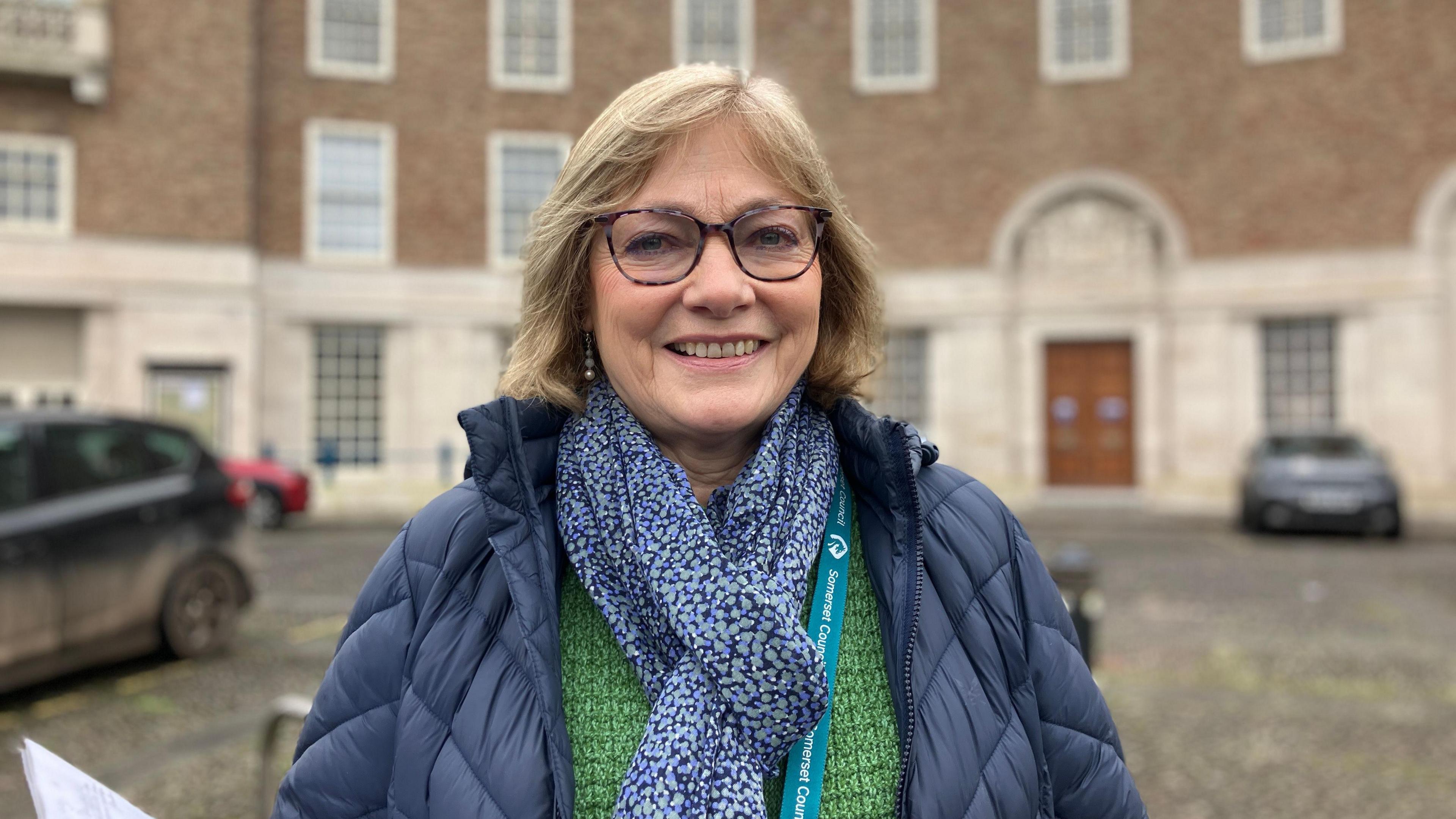 Sarah Wakefield  is standing in front of the council building. She is wearing a blue scarf and a blue jacket. She is smiling at the camera.