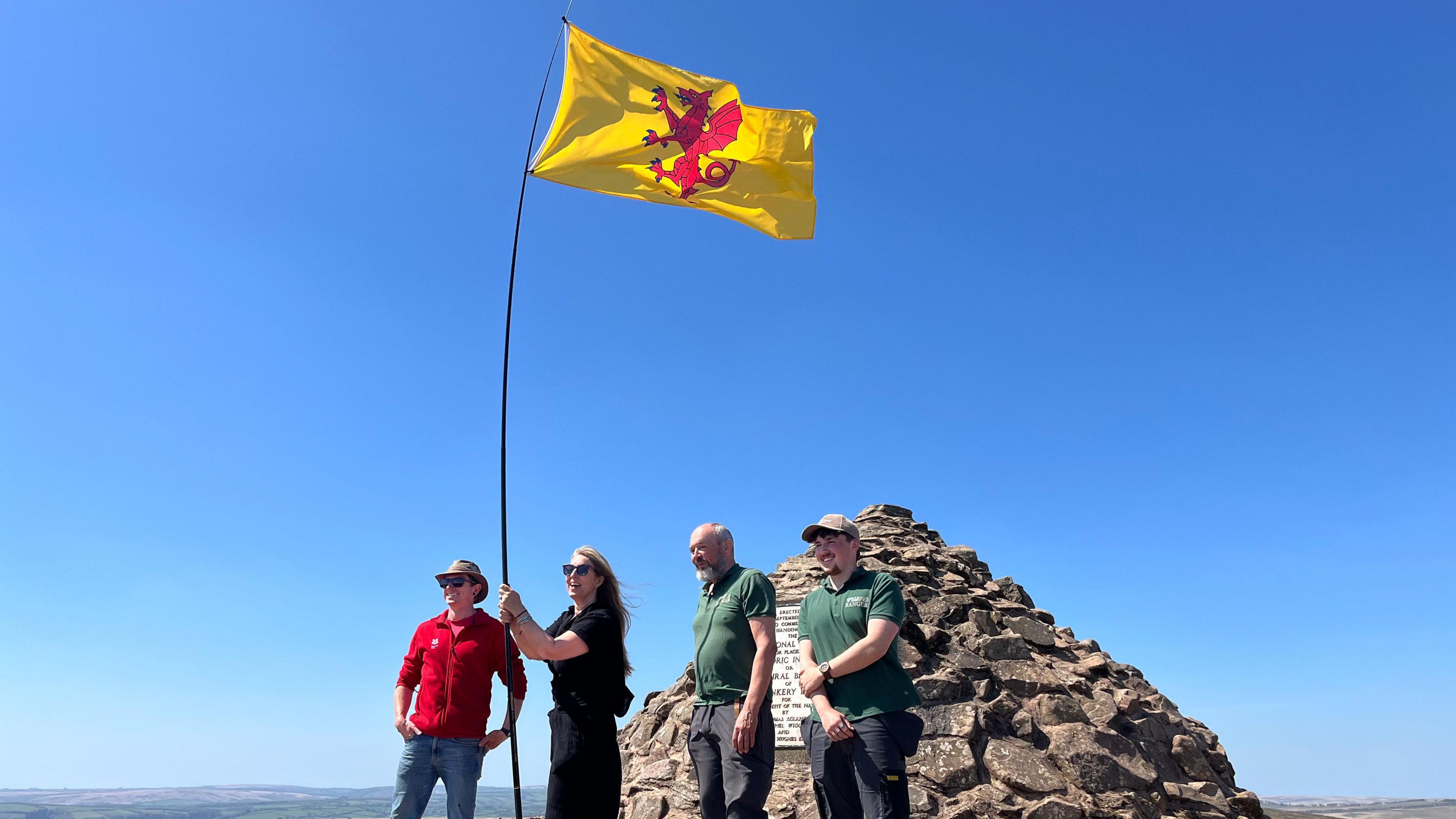 Four people are standing on the top of a mountain holding up a long pole with the Somerset flag on the end.