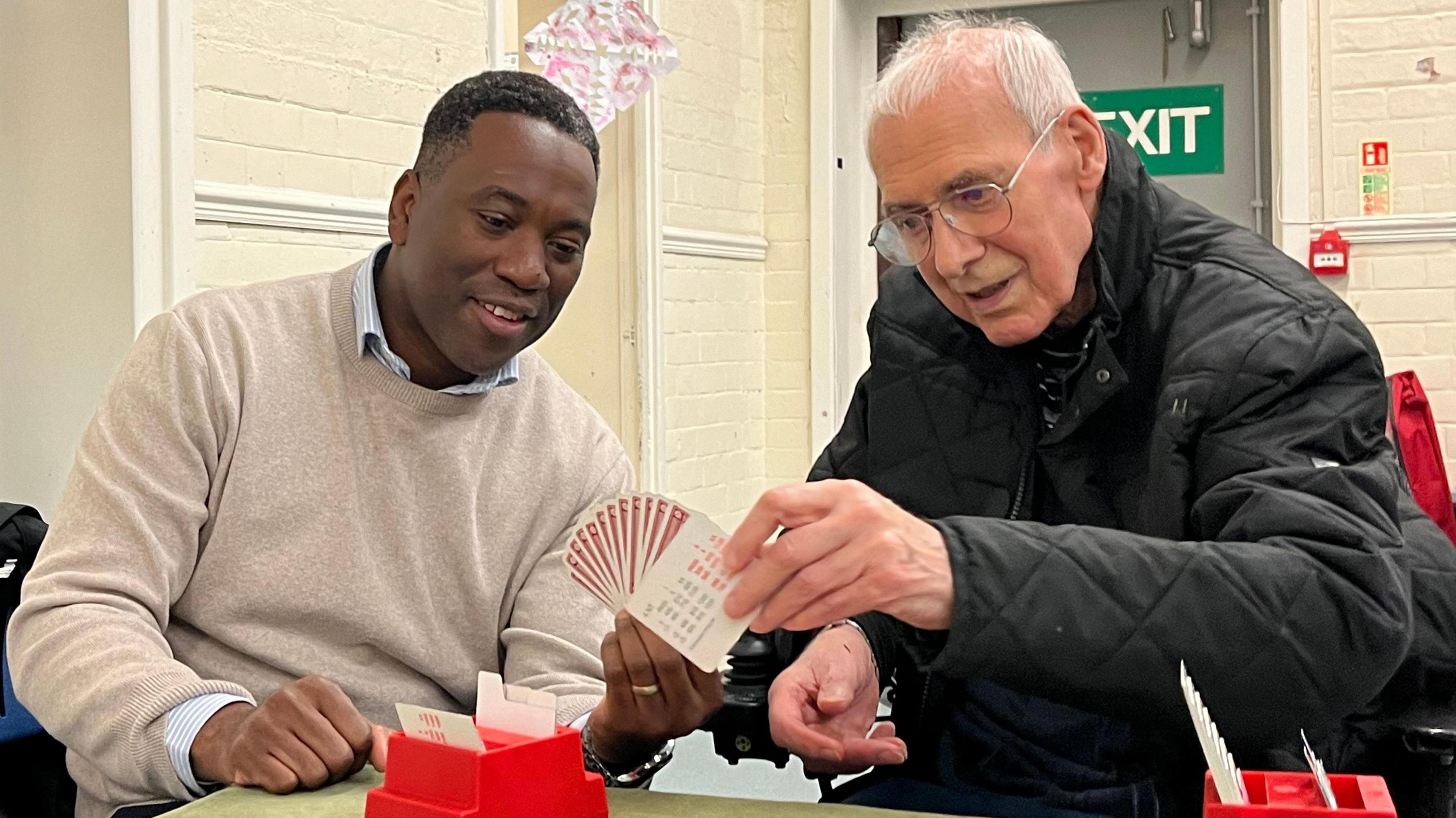 Bright and Peter sit next to each other at a bridge table, Bright is holding the hand of cards for Peter to look at. Peter is choosing a card to put down from the hand. The table has a green velvet cover and there's red boxes holding the bidding cards. 