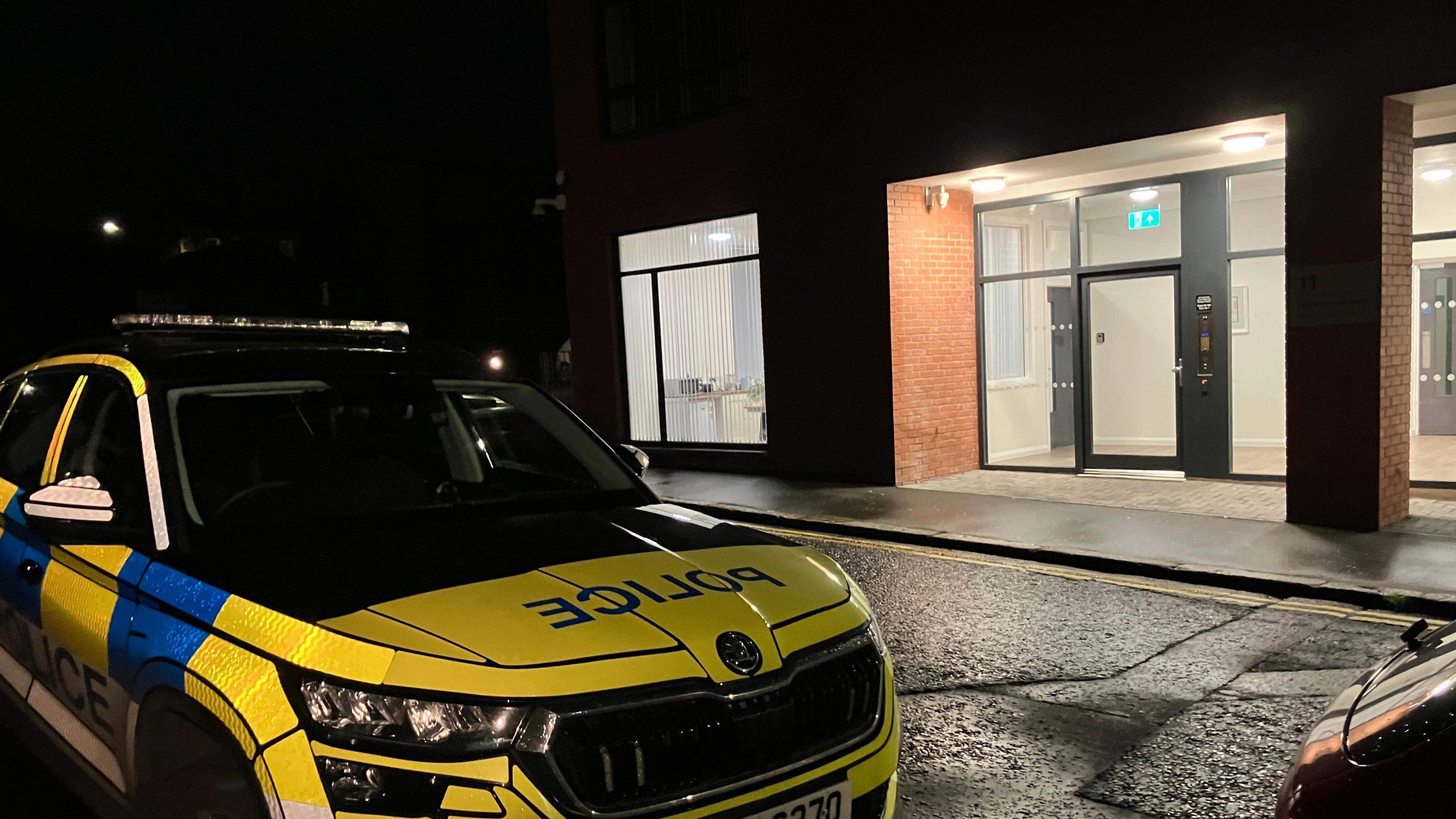 A police car outside an apartment block in Lisburn. Only the ground floor of the red brick building is visible in the photo. There are large windows around the front door and lights are on inside the building. The police car is yellow, blue and white and has the word "police" on the bonnet and side.