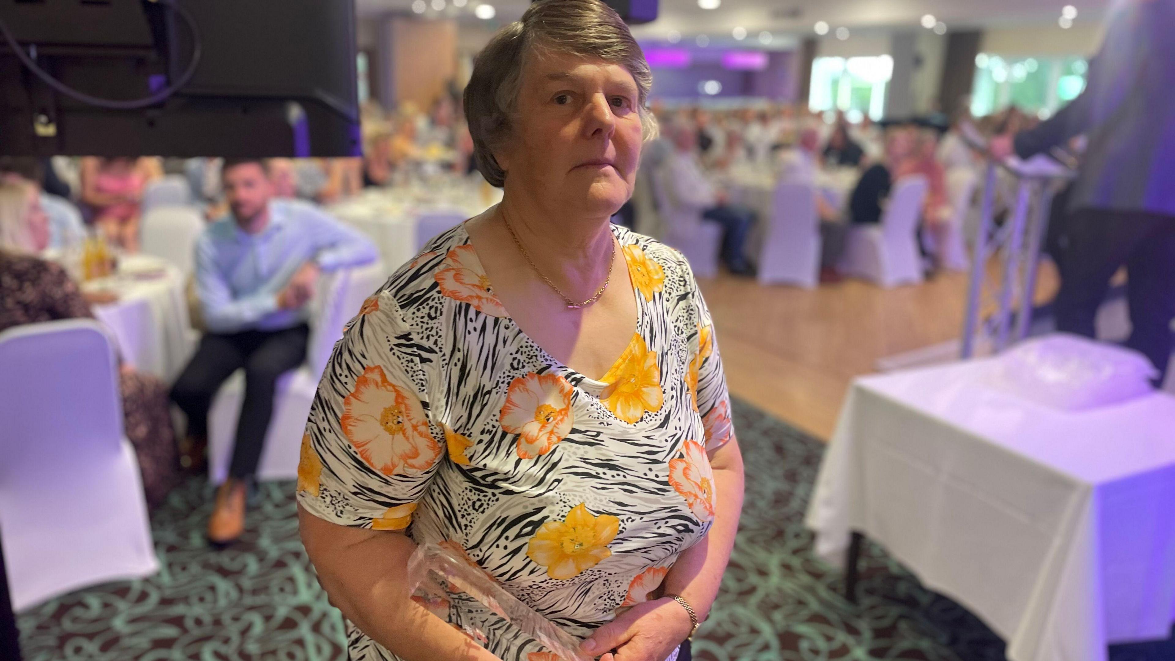 Margaret Grieve standing with her great neighbour award at the ceremony. There are people in the background sitting at tables.