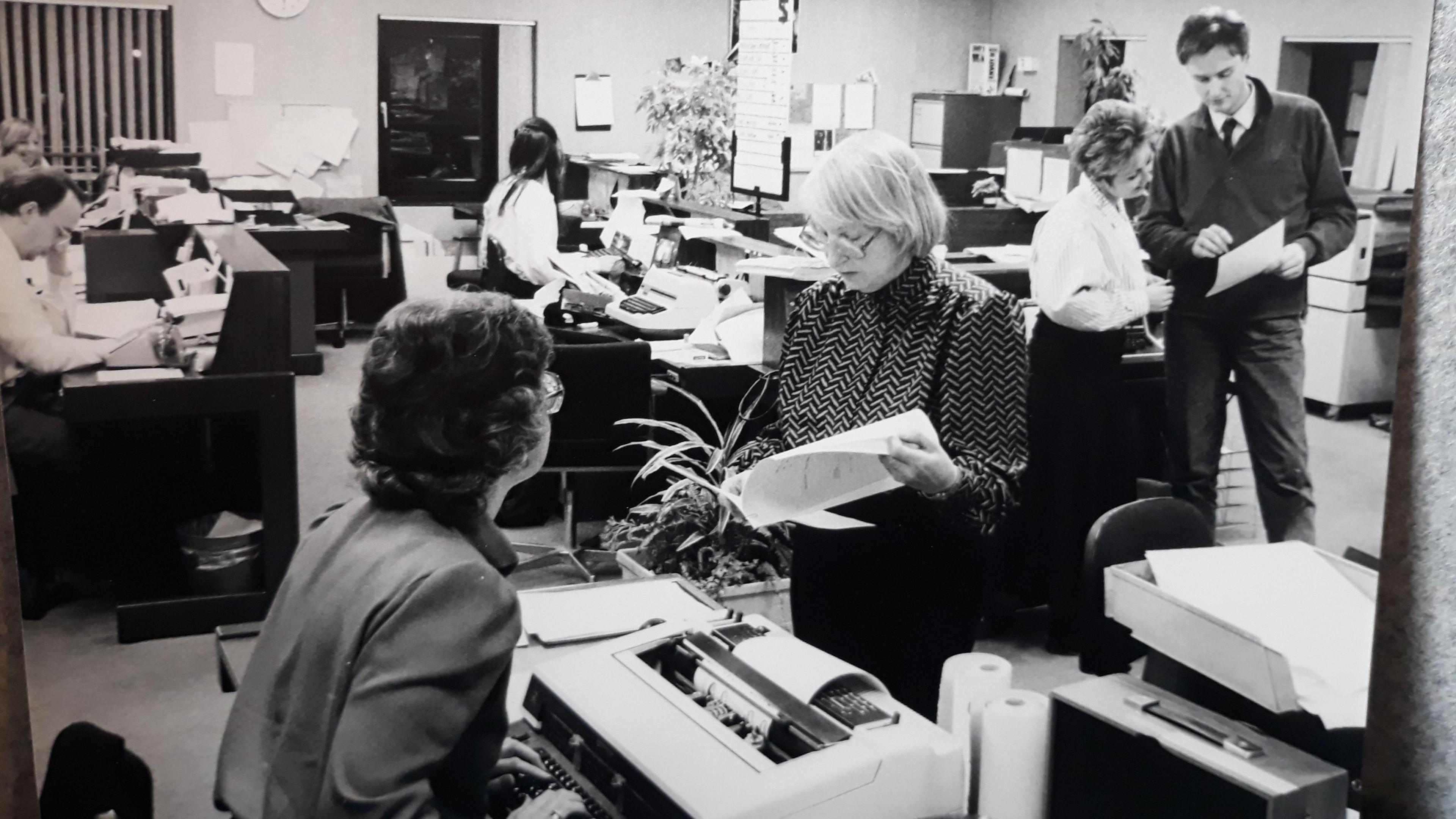 People working in the Look East newsroom in 1987. Several people are seated at desks with typewriters. A man and woman are standing up, looking at a piece of paper and talking.