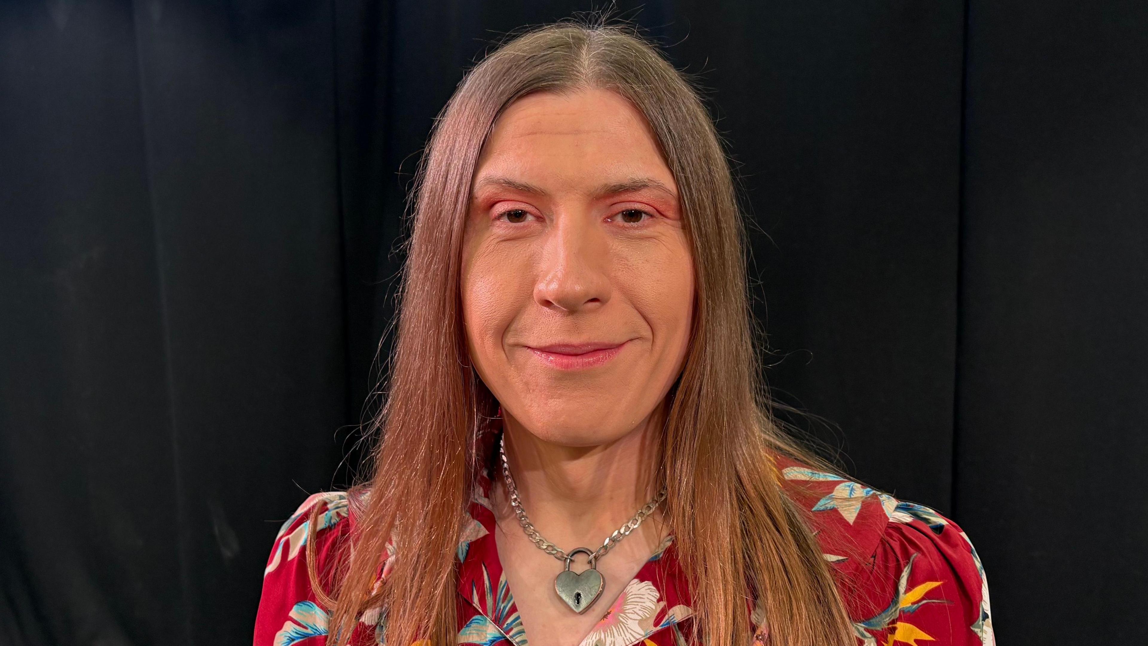 Jessica Brown looking directly at the camera. She has straight brown hair, which falls just below her shoulders. She is wearing a red dress with large flowers on it, though only her head and shoulders are in the frame. Around her neck is a silver chain with a heart-shaped padlock pendant. She is smiling at the camera and is sitting in front of a black backdrop.