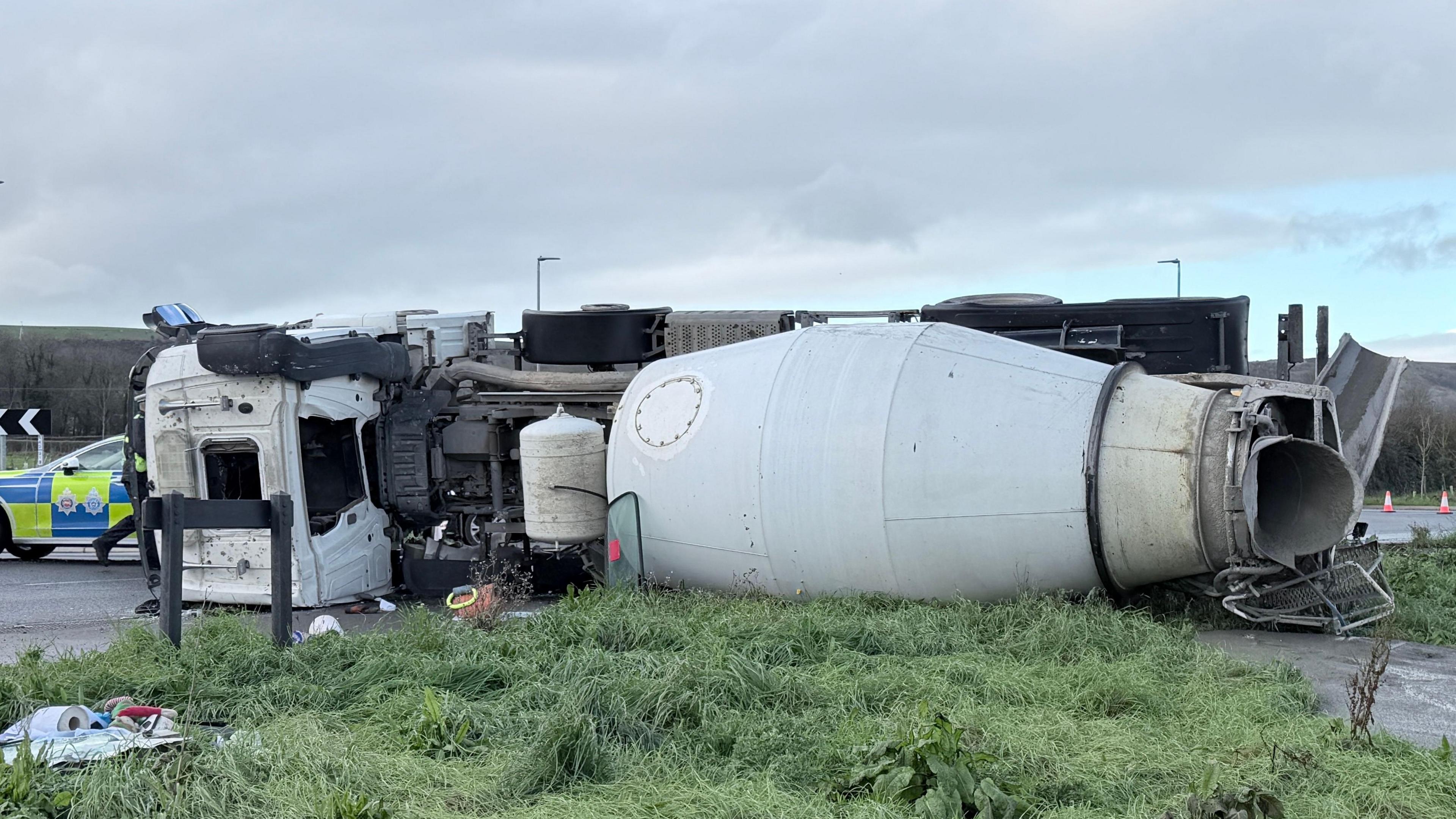 The image shows a close-up of the overturned HGV lying on the grass, with a police car visible in the back on the left-hand-side. The sky is a light blue with grey clouds.