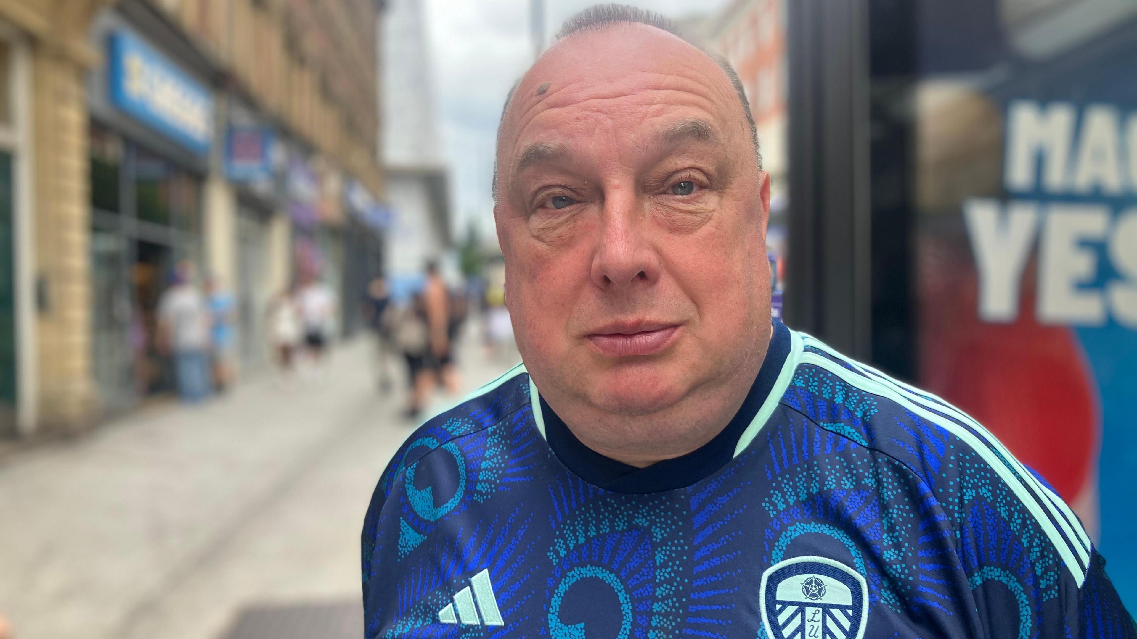 A man wears a blue Leeds United Football Club shirt. Behind him, Leeds city centre is blurred.