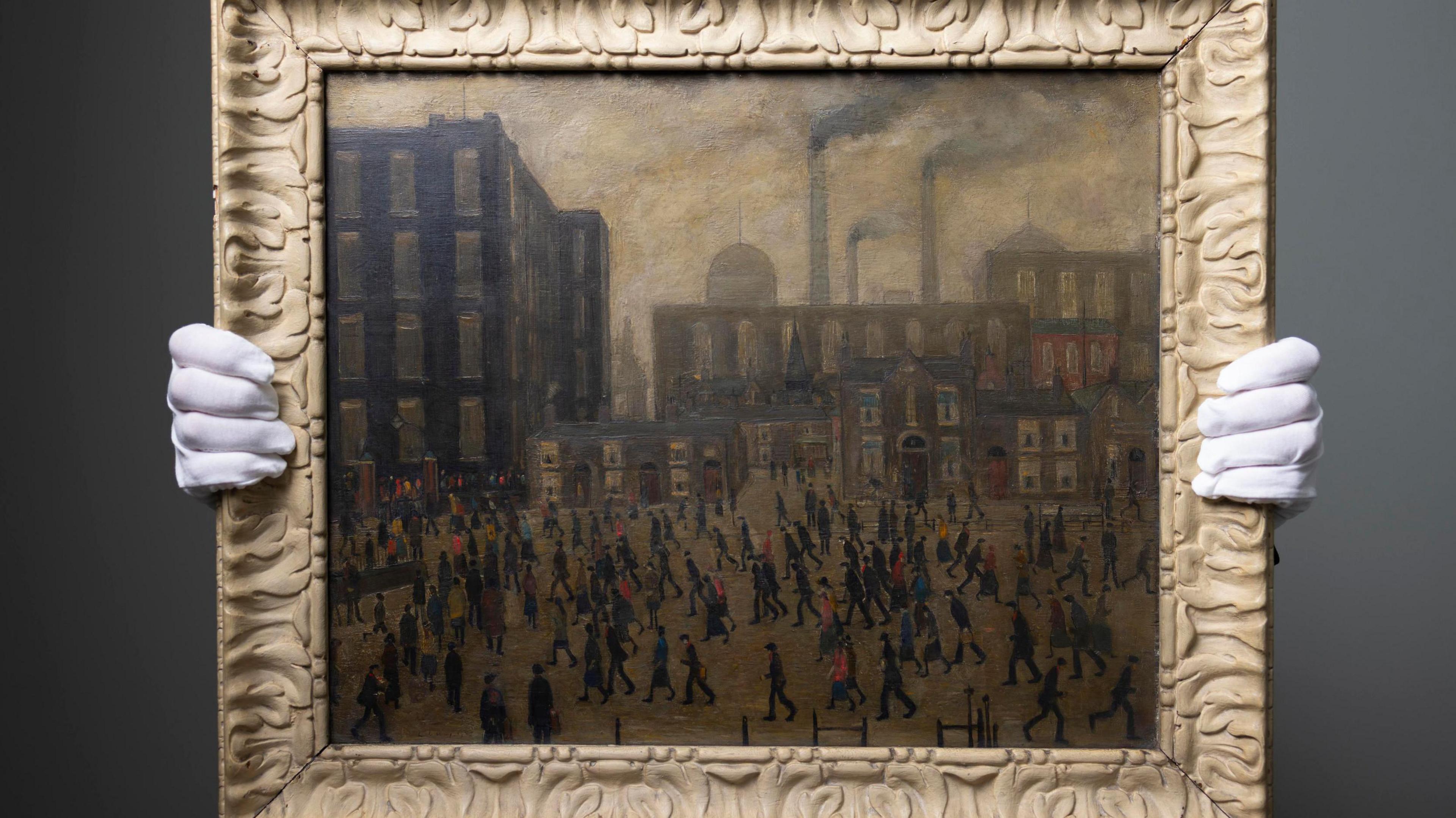 Someone with white gloves holding a framed edition of one of L. S. Lowry's paintings. It has a dark gloomy colour palette. Hundreds of people are walking across the scene and factories are pluming smoke from chimneys in the background.
