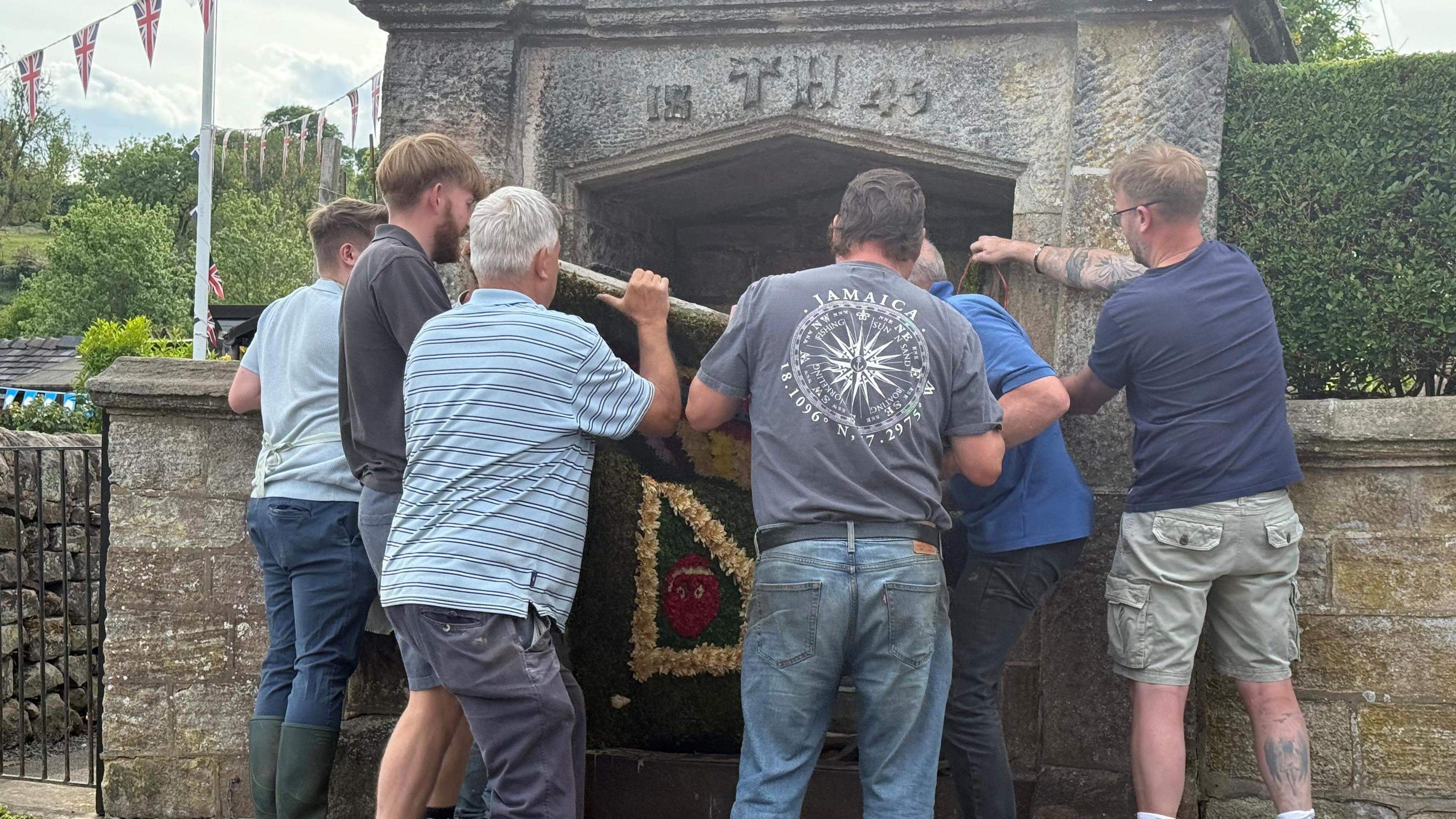 A group of people lift a decorative board onto a stone well. There is a gate to the side and a hedge on the other side of the well.