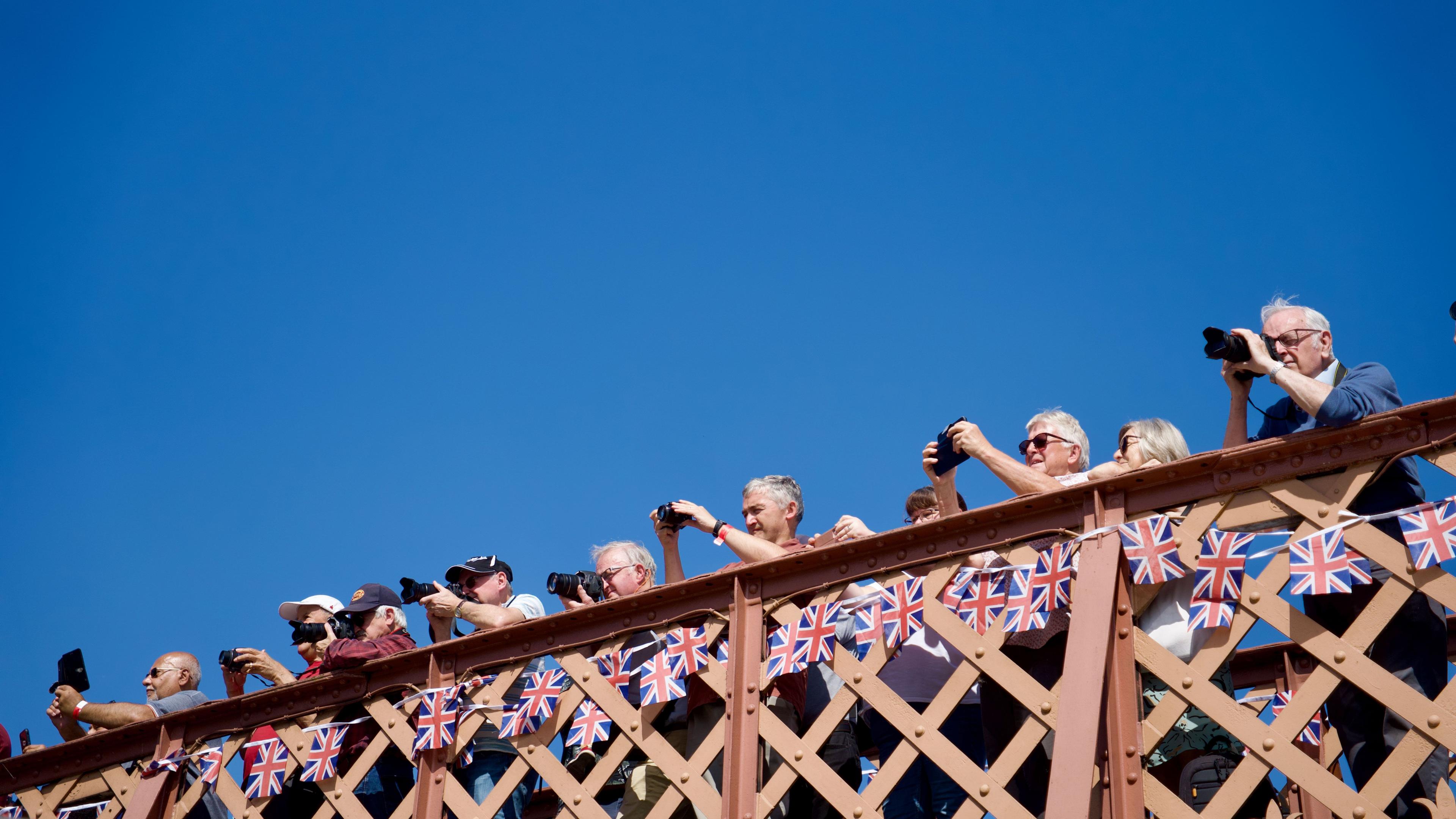 Ten people on a bridge over a railway taking photographs. There is union jack bunting hanging from the bridge