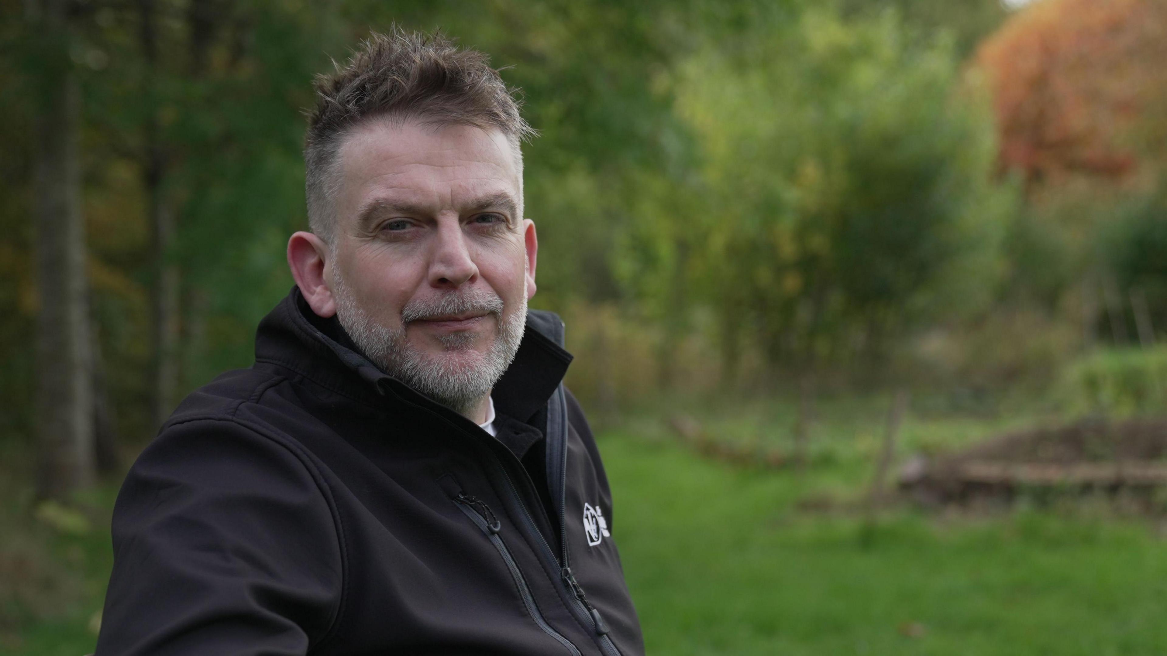 Dom wears a black fleece jacket with the Wildlife Trusts log while sitting on a bench in a field, surrounded by trees