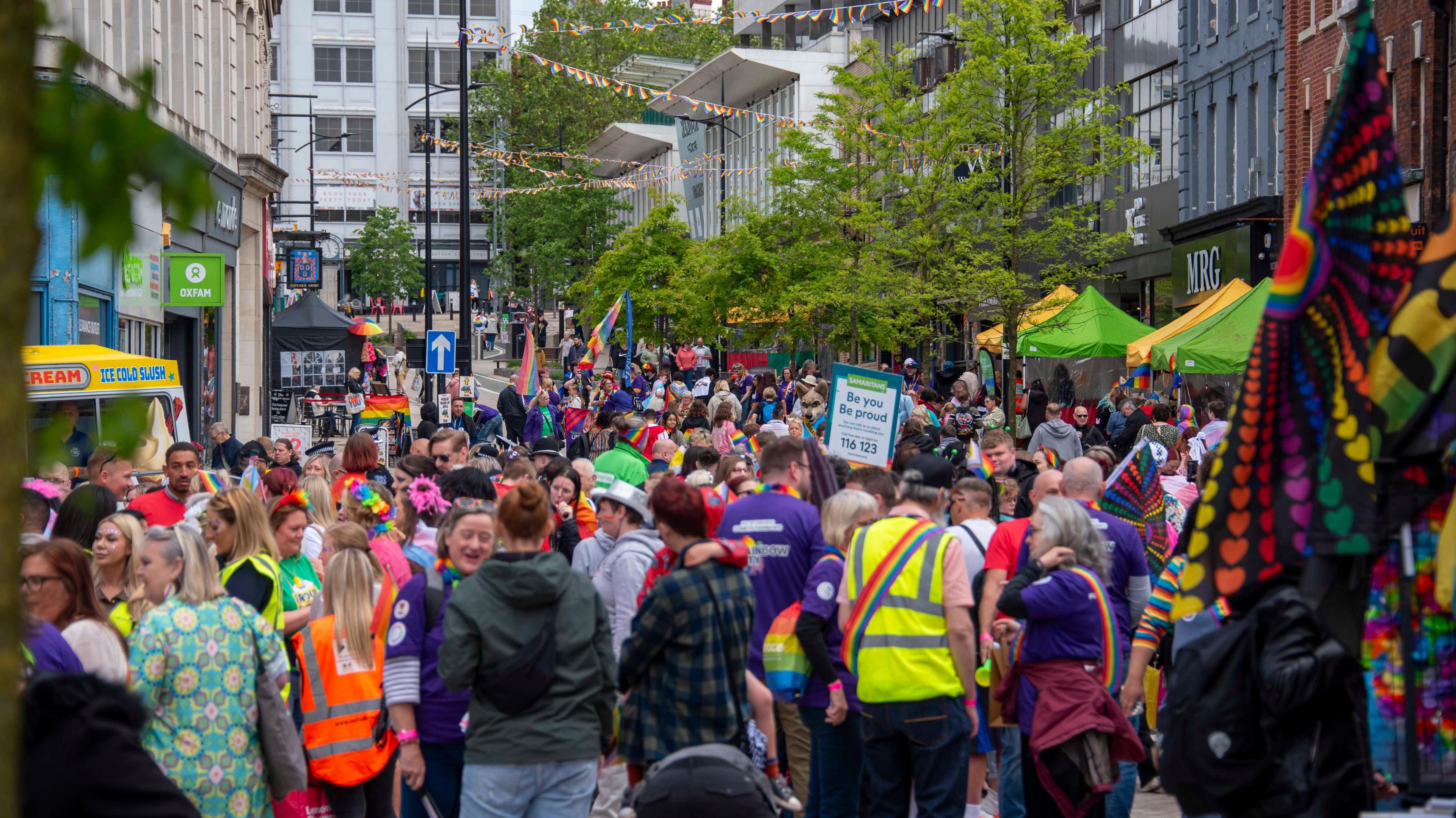 A crowded city centre street with brightly coloured bunting. Some people are carrying Pride flags, some are carrying banners, most are wearing bright coloured clothes