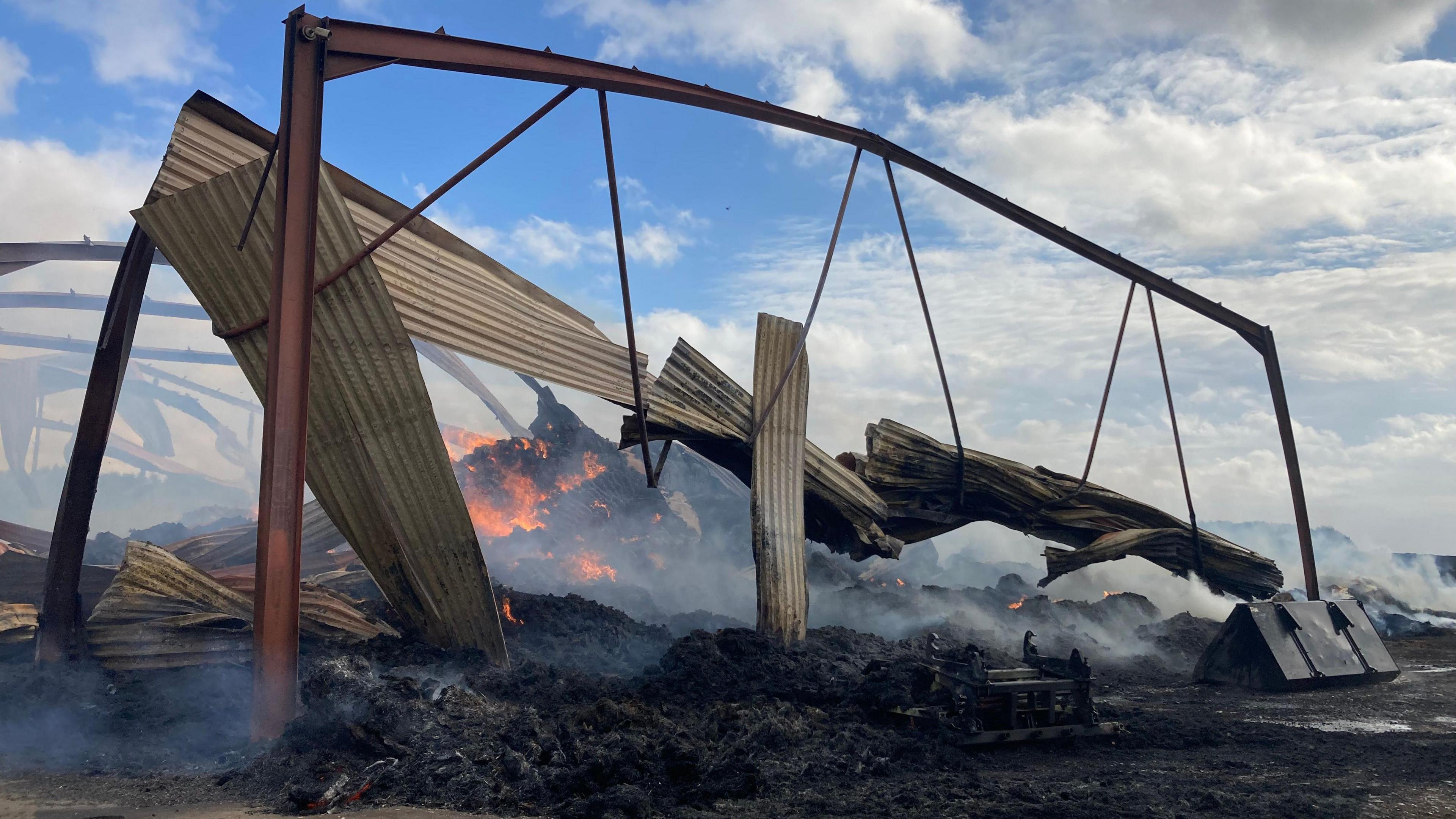 The barn which has been completely destroyed by the fire. The metal frame of the building is barely standing, with a pile of rubble on fire in the centre of the remains. The ground is smouldering.
