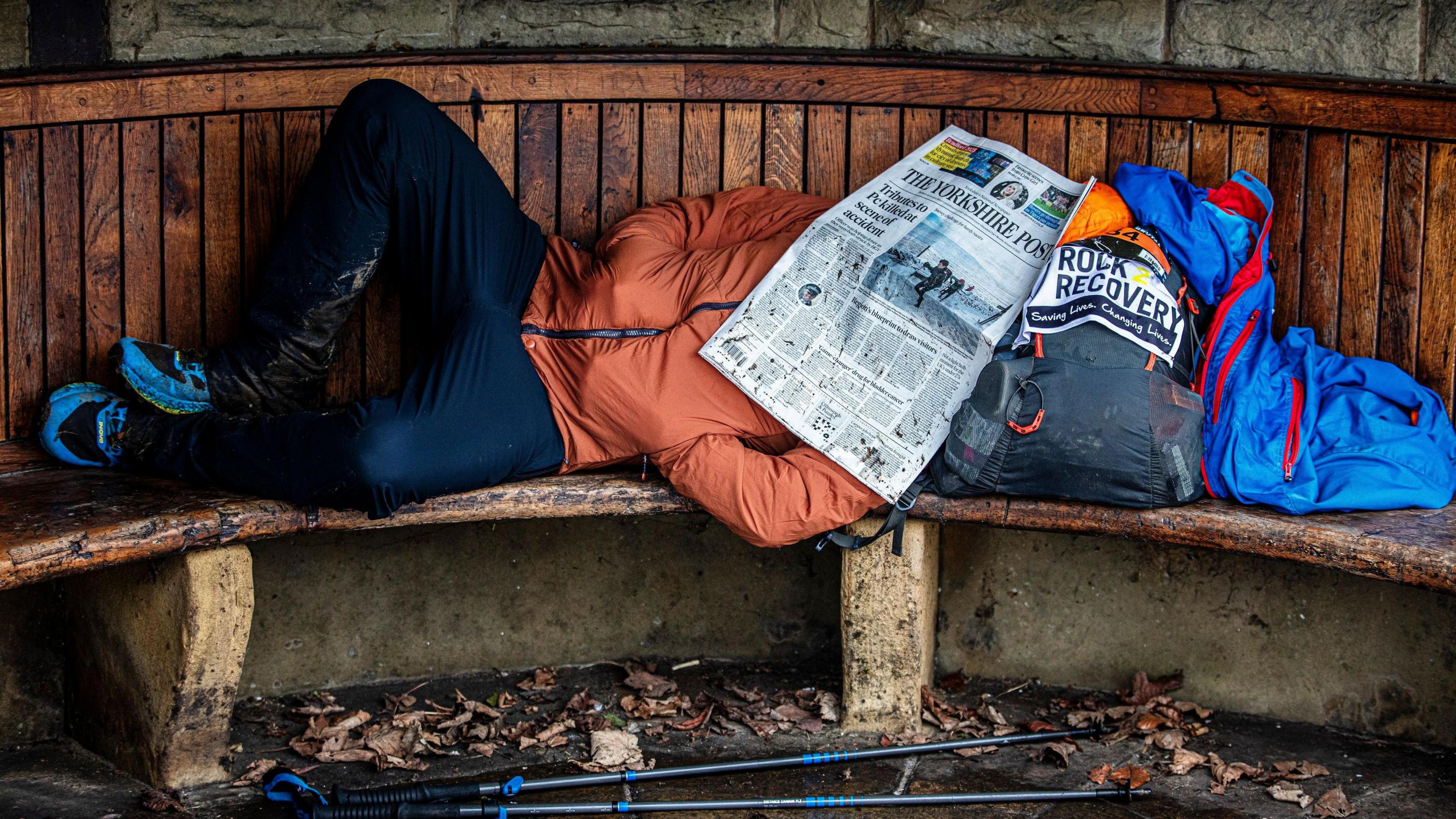 A race, wearing blue leggings and an orange jacket, lying on a bench with a rucksack for a pillow and a newspaper over his face