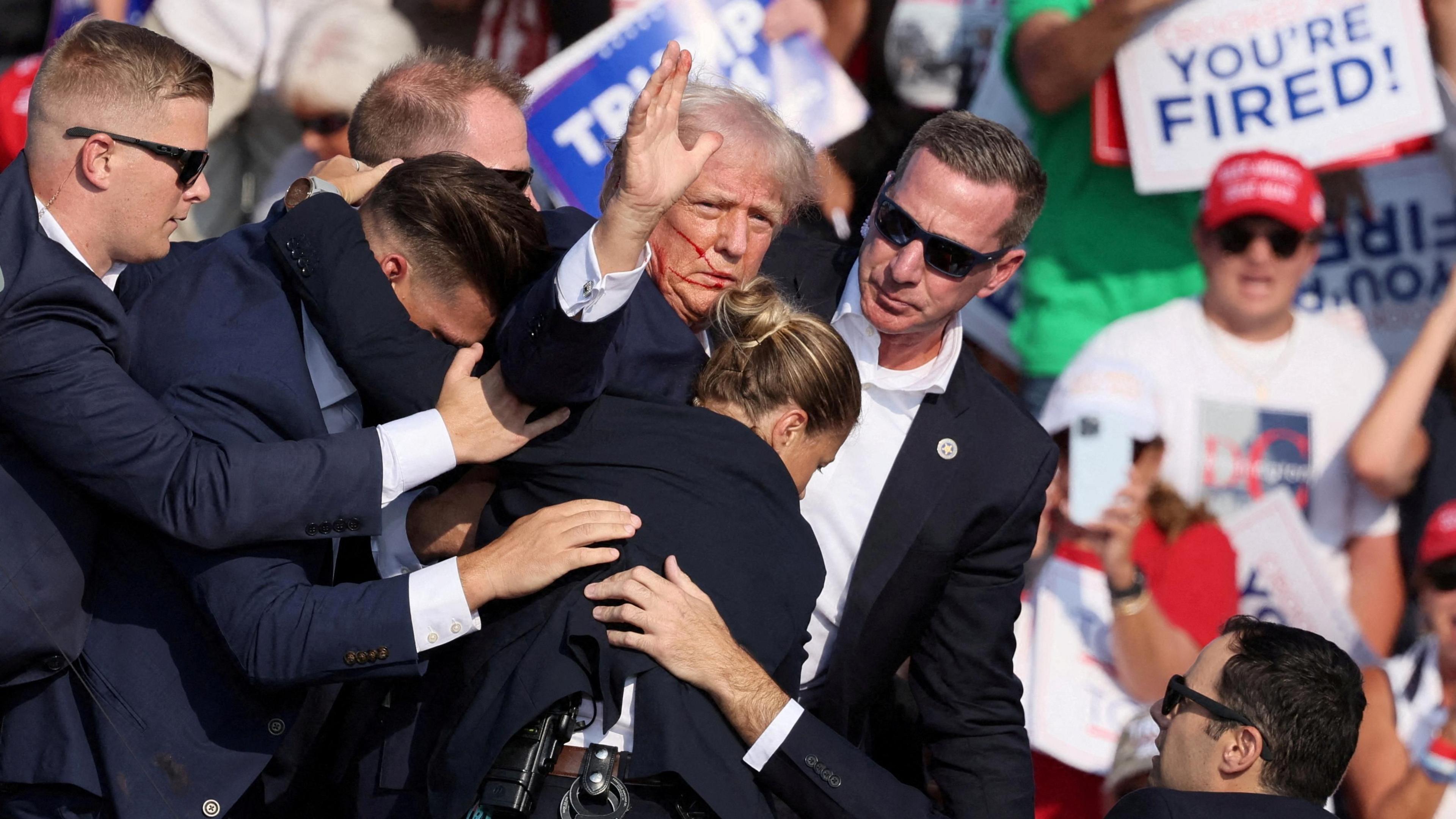 Secret service bodyguards crowd around a waving Donald Trump and try to usher him off stage after he was shot at during a rally in Butler, Pennsylvania on July 13