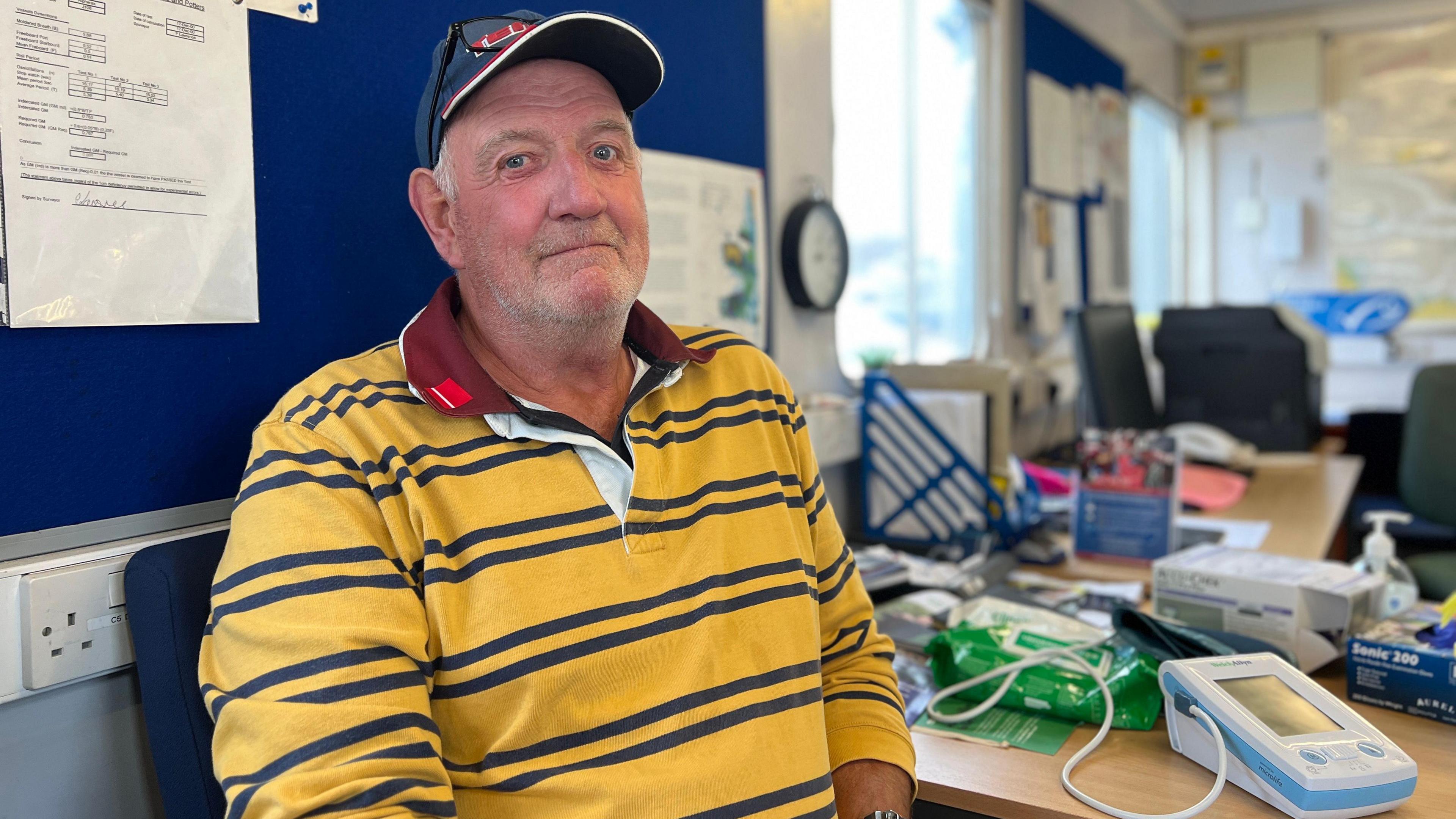 Chris looks at the camera as he's sat by a desk which has medical equipment to measure his cholesterol. He is wearing a yellow jumper with blue stripes and a red collar. He has a blue cap on with glasses on the rim of the cap. Behind him is a blue board with a few lamentated pieces of paper pinned to it.