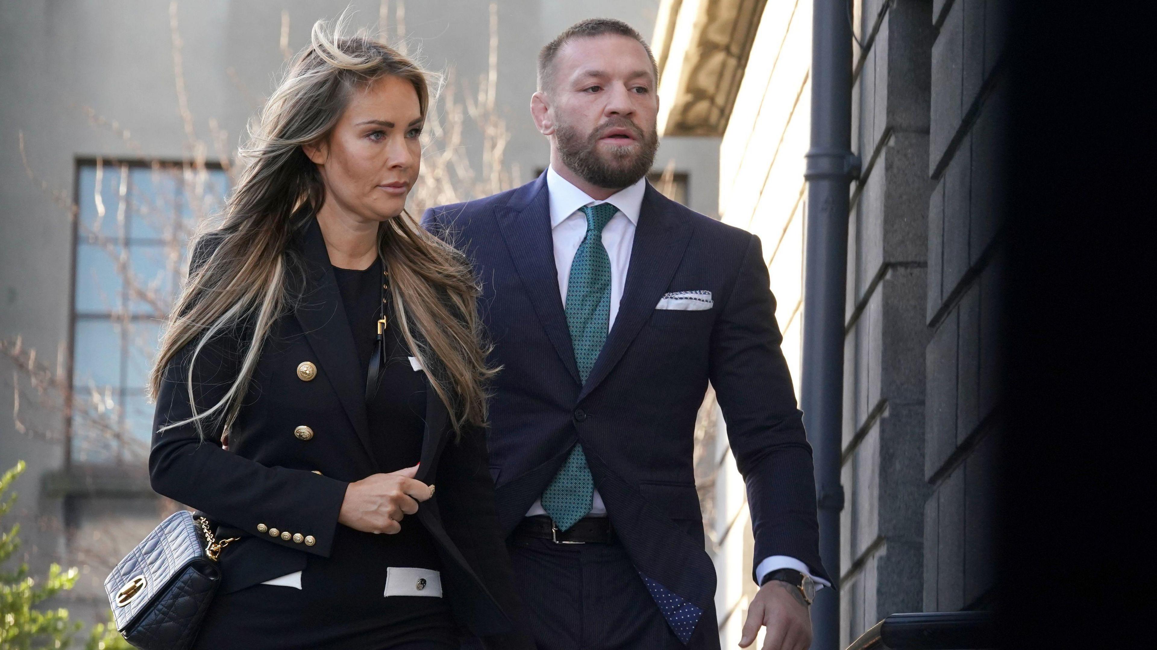 Pictured outside a grey building, are Conor McGregor, wearing a navy suit, green tie and white shirt, beside his partner Dee Devlin, a woman with long blonde hair, wearing a black top and black blazer. 