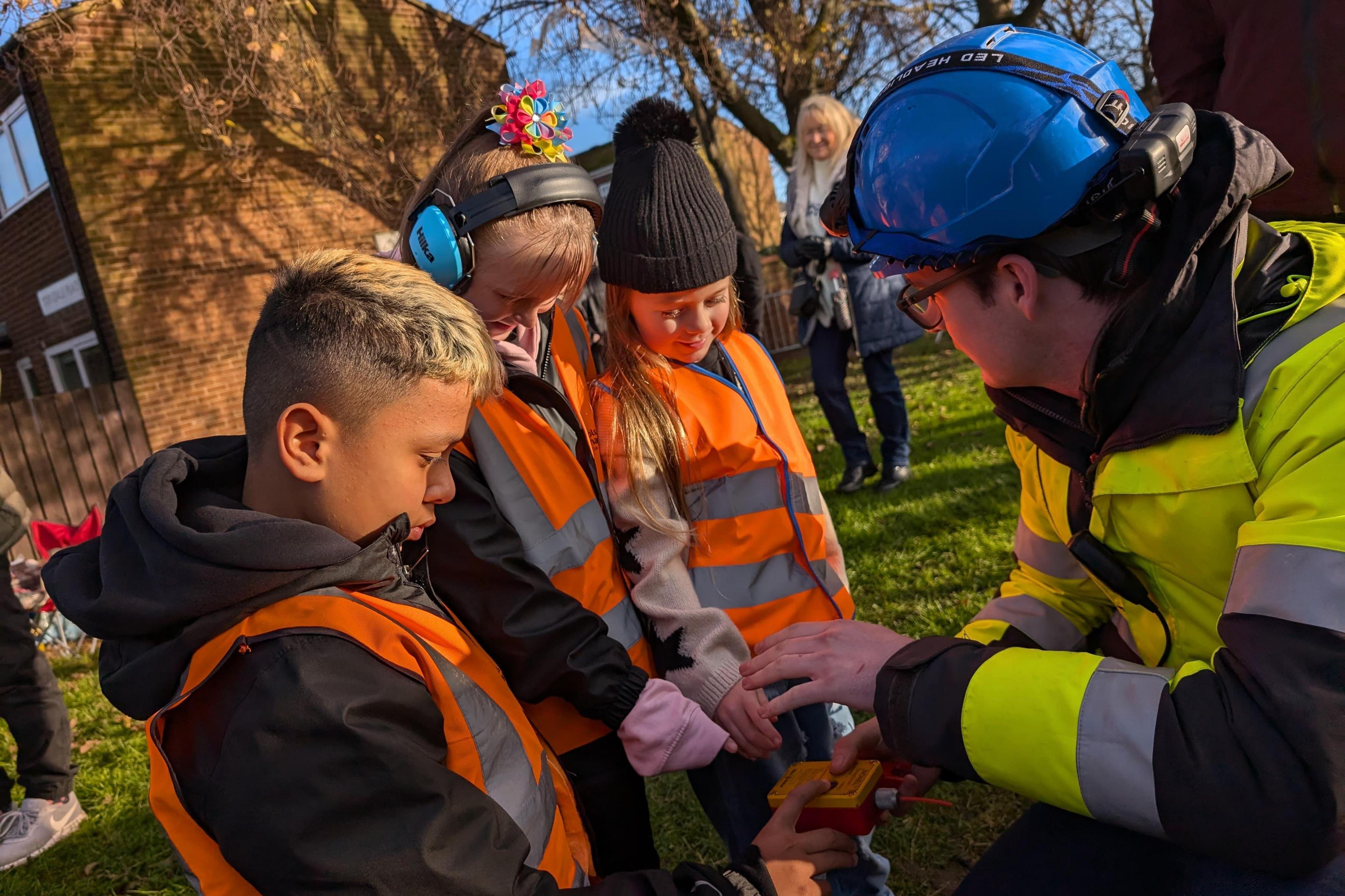 Children - Aidan, Emily-Jane and Frankie, are shown the demolition button by Cormac. They are wearing high-vis jackets.