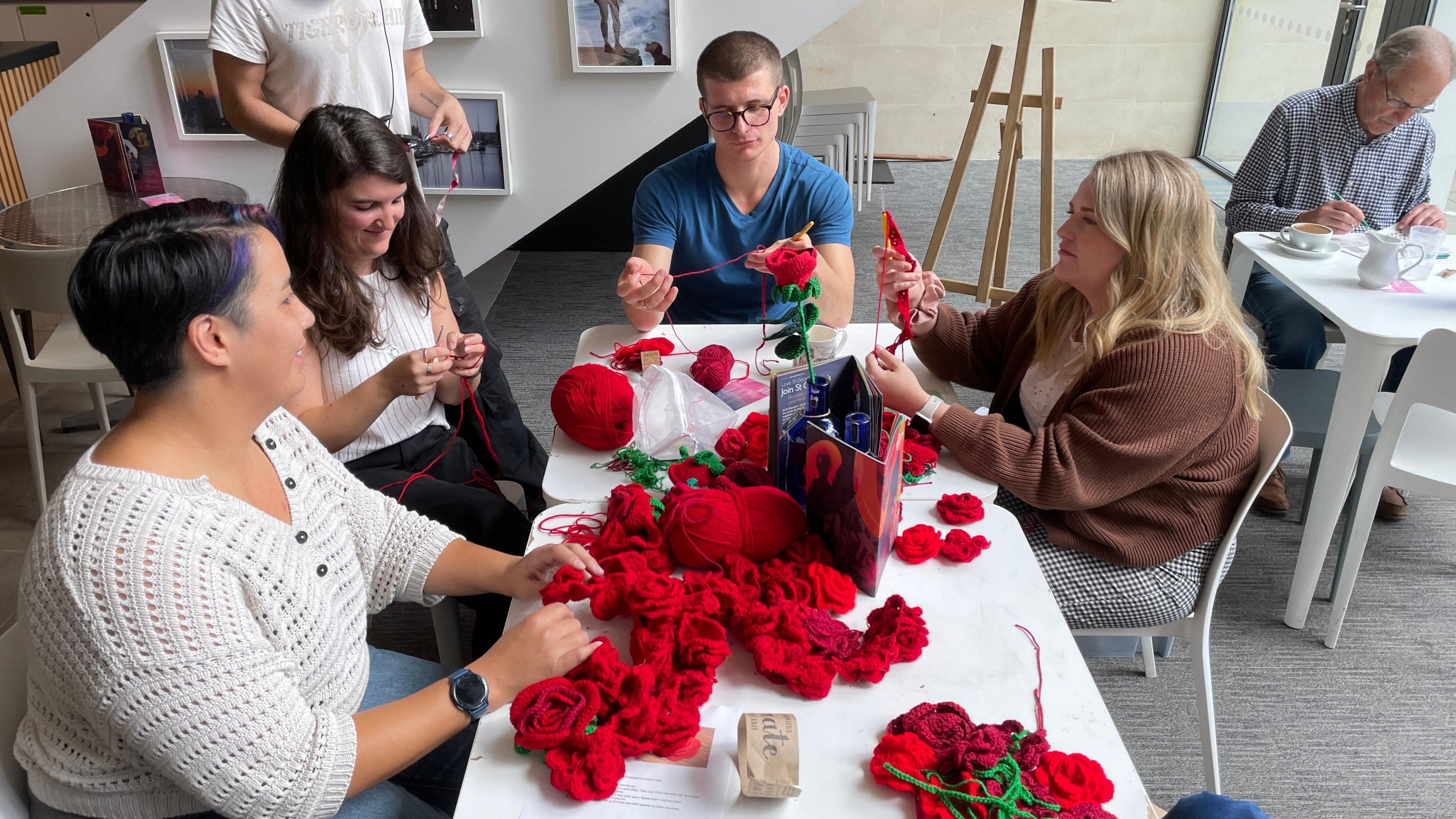 A group of people sitting around a long table crocheting lots of red roses. There is a large pile of them in the centre of the table, and balls of yarn further down.