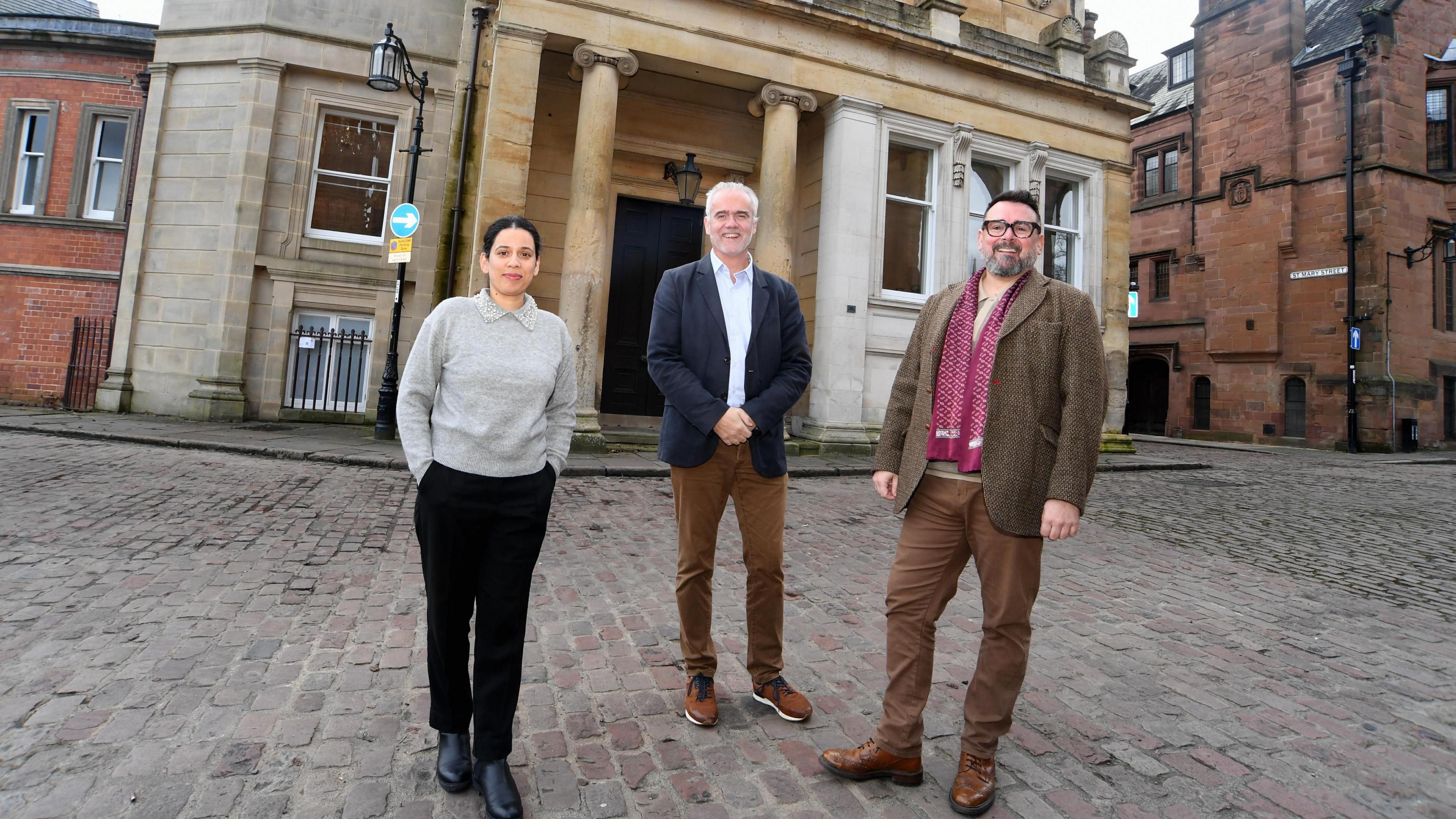 Three people stand in front of Drapers' Hall in Coventry. There is a woman with dark hair tied back, wearing a grey pullover, black trousers and black shoes. There is a man with short white hair wearing a dark blue jacket, a light blue shirt, brown trousers and brown shoes. A second man with short dark hair, glasses and a grey beard is wearing a brown jacket, brown trousers and brown shoes with a burgundy scarf.