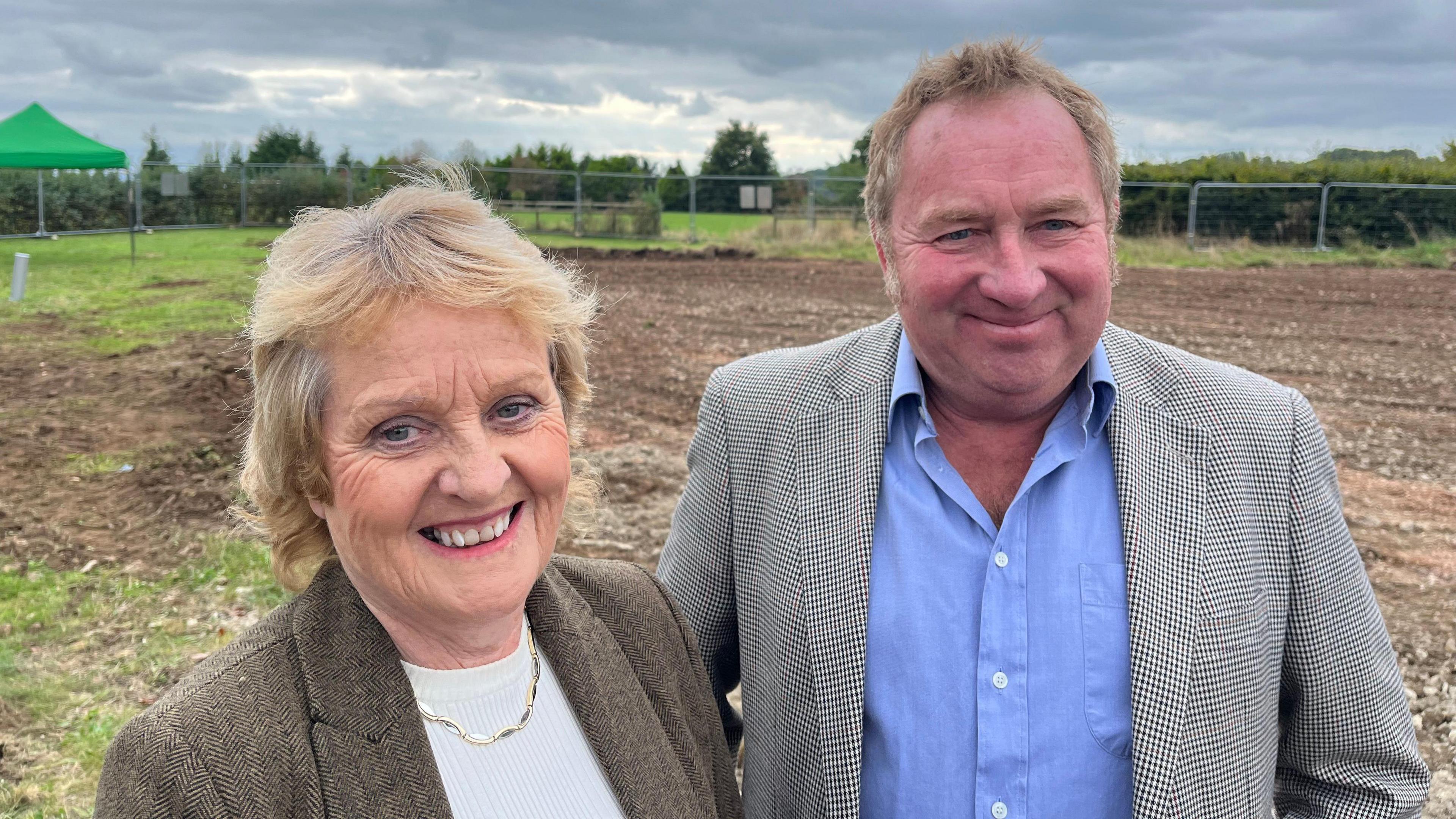 Sue Wilkinson standing next to Jim Butler. They are wearing blazers and smiling. They stand in an empty field, where the village hall will be built.