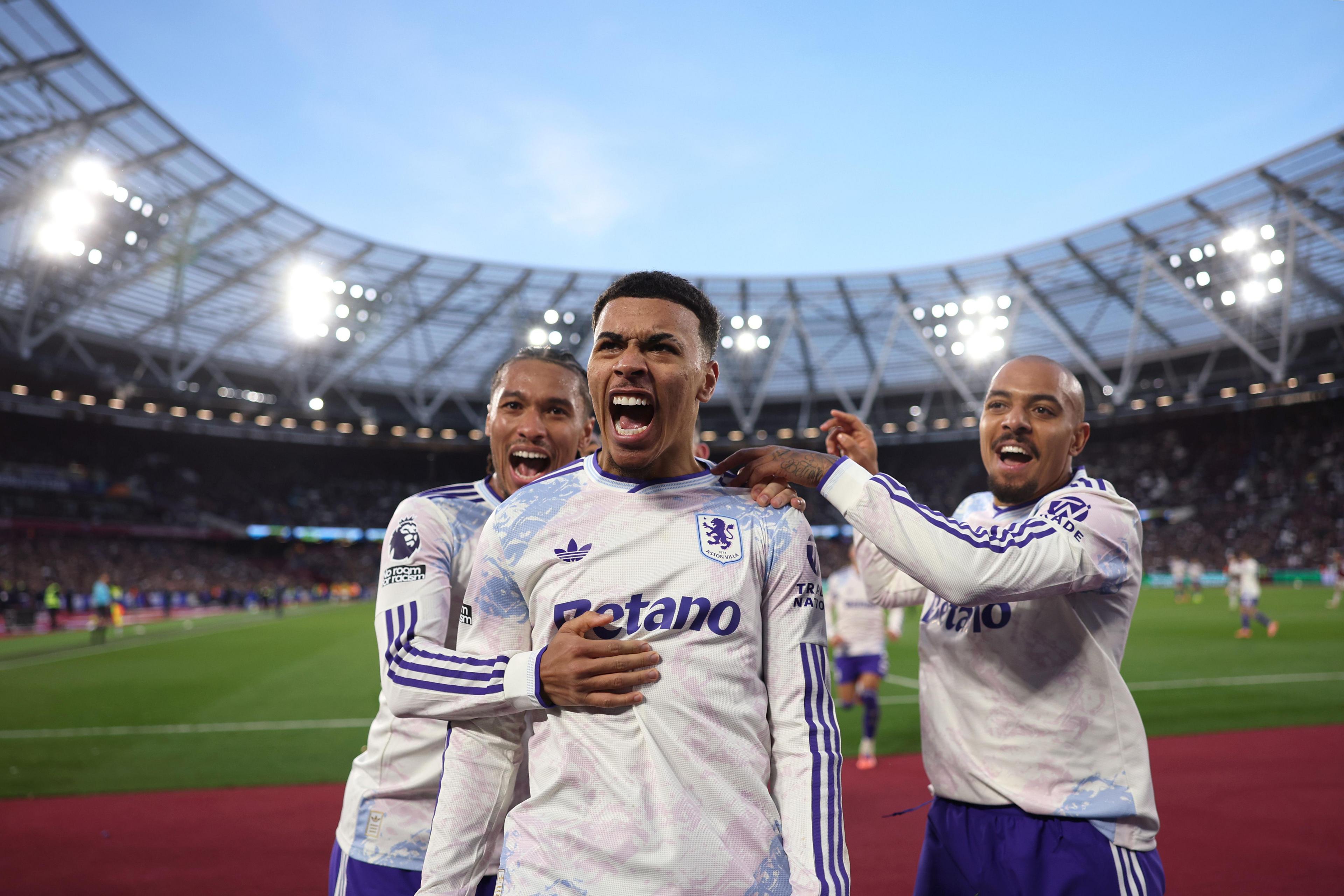 Tres jugadores de fútbol celebran juntos cerca de la línea de banda de un estadio, mientras la multitud observa desde las gradas bajo las brillantes luces del estadio.