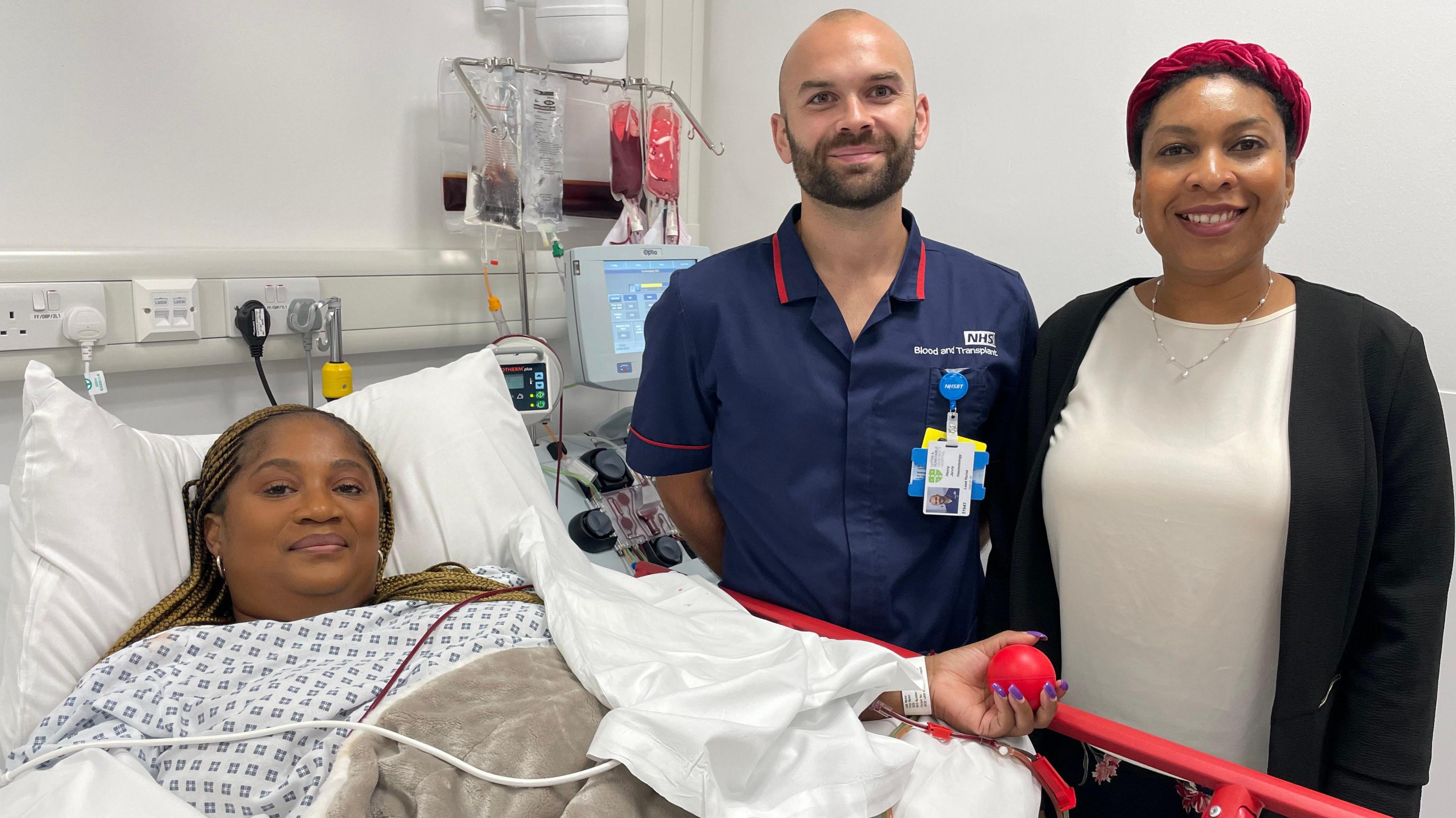 A woman is laying in a bed with a man wearing a nurse uniform stood next to the bed and a woman smartly dressed next to him.