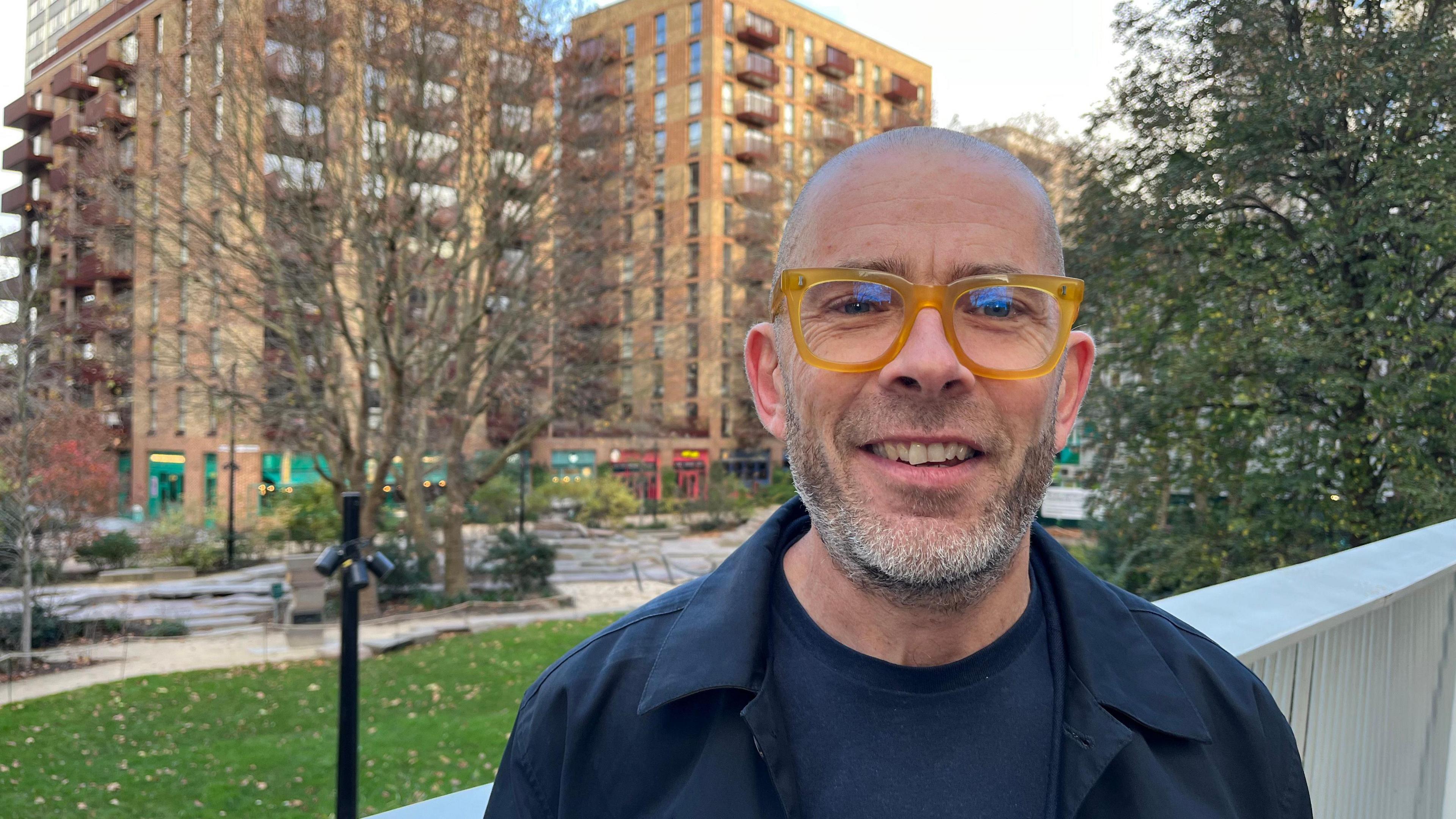 A man in glasses stands in front of a block of flats 