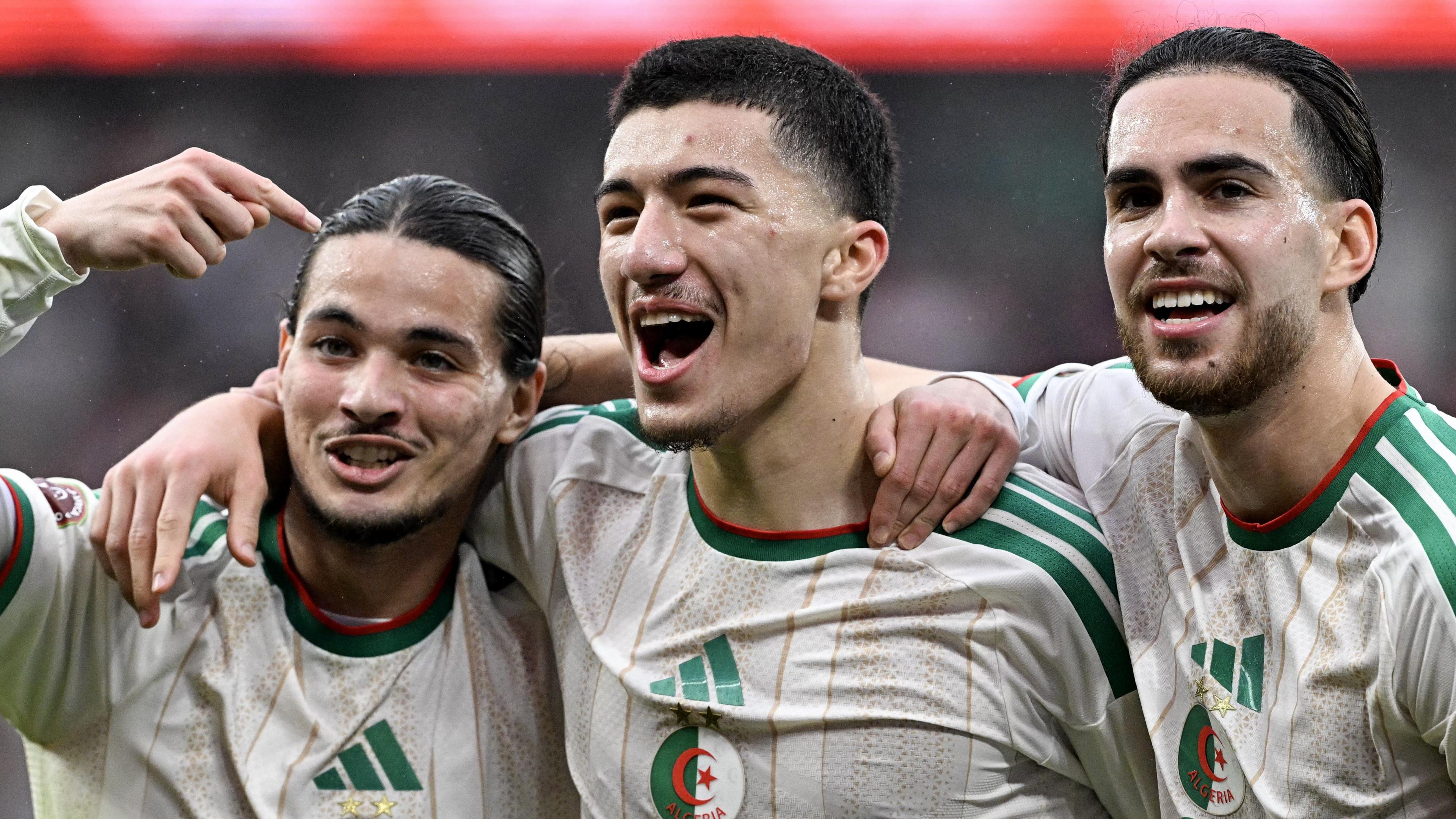 Algeria trio Anis Hadj Moussa (left), Ibrahim Maza (centre) and Ramiz Zerrouki (right) stand in a line to celebrate scoring their team's third goal during the 2025 Africa Cup of Nations game against Equatorial Guinea. The players are all visible from chest up in white Algeria away jerseys with green and gold trim. Moussa is using his right hand to point back along the line at his team-mates