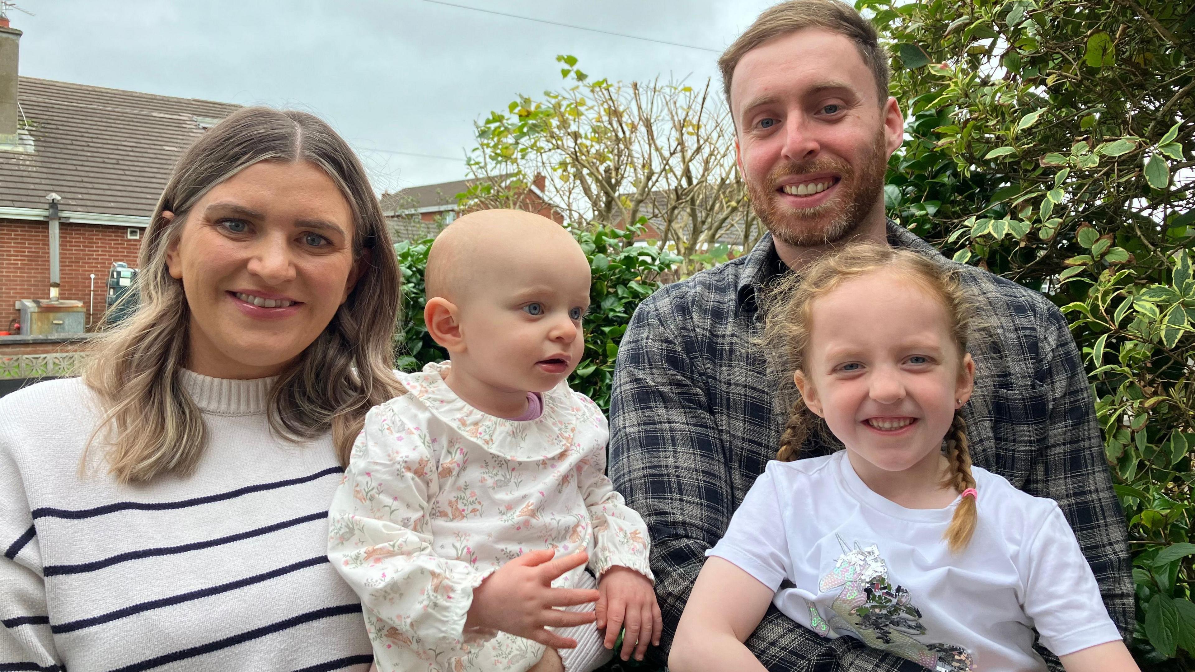 Craig and Anna are standing next to each other in a garden with a bush and a house in the background. They are smiling into the camera. Lauren is holding Anna and Craig is holding their other daughter.