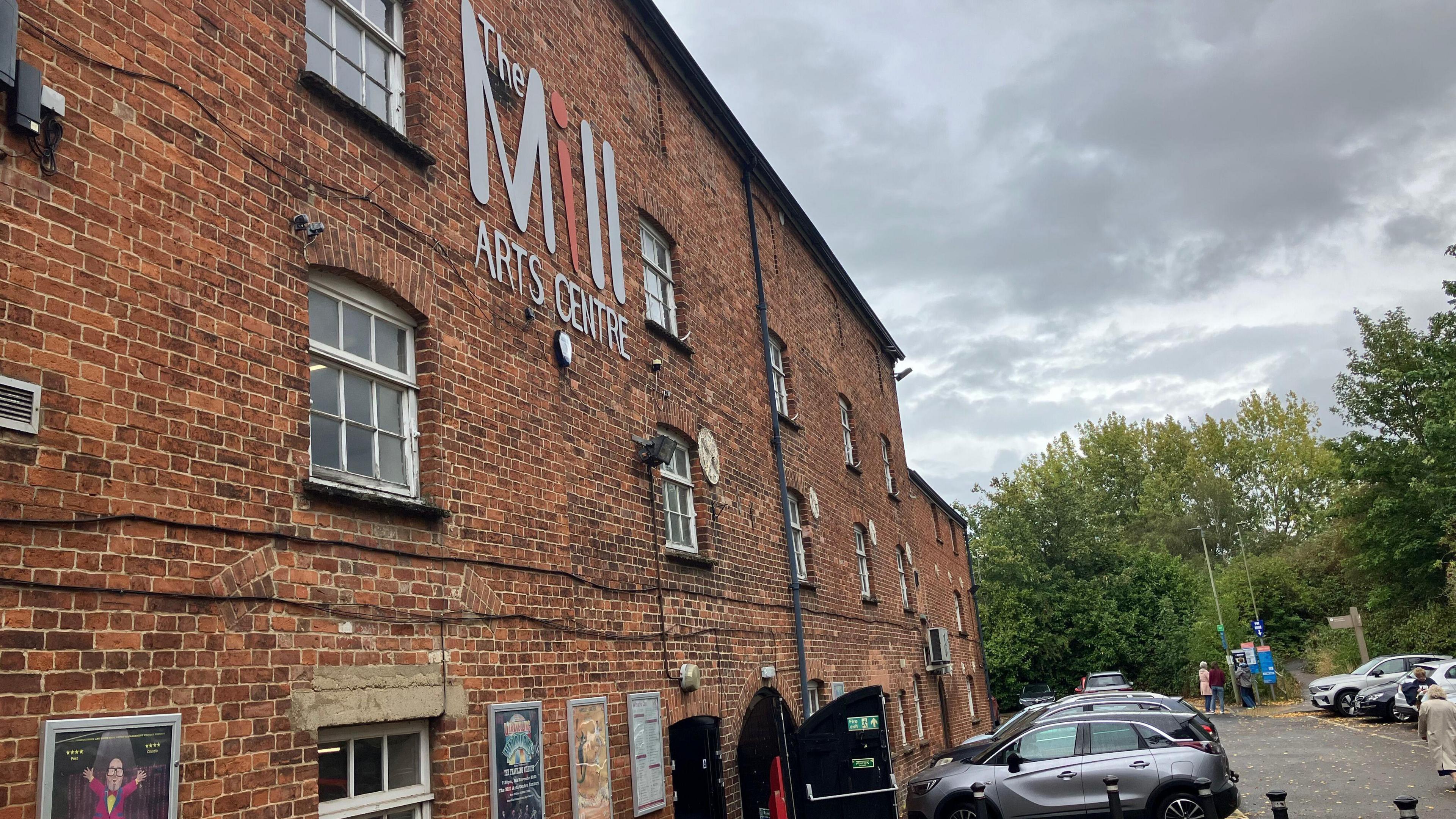 The outside of the Mill Arts Centre. It is a Victorian-style red brick building, with a large sign saying "The Mill Arts Centre" on the front.