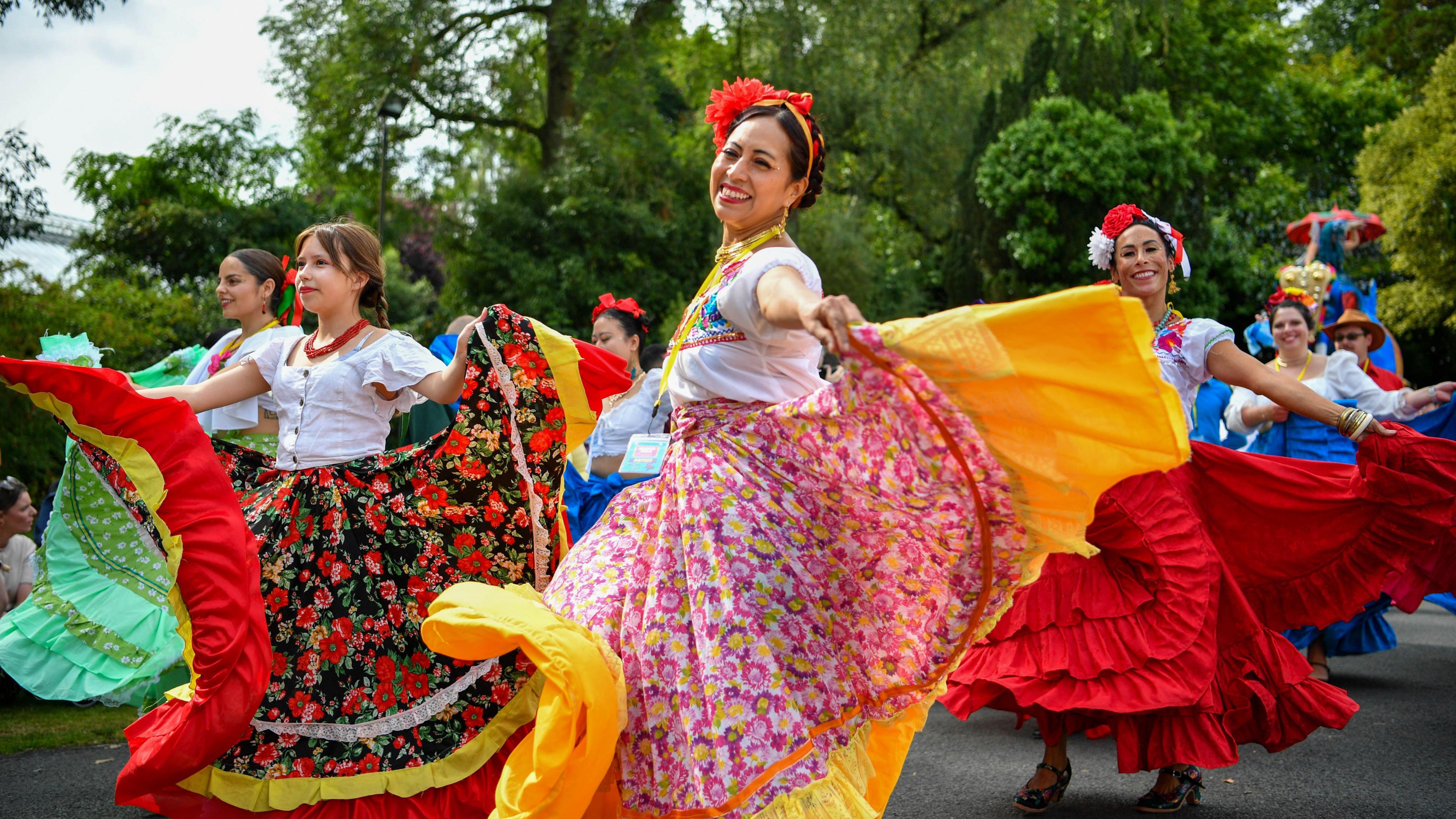 Women are dressed in colourful dresses ad dancing in a festival. Some have flowers in their hairs. Trees are behind them. 