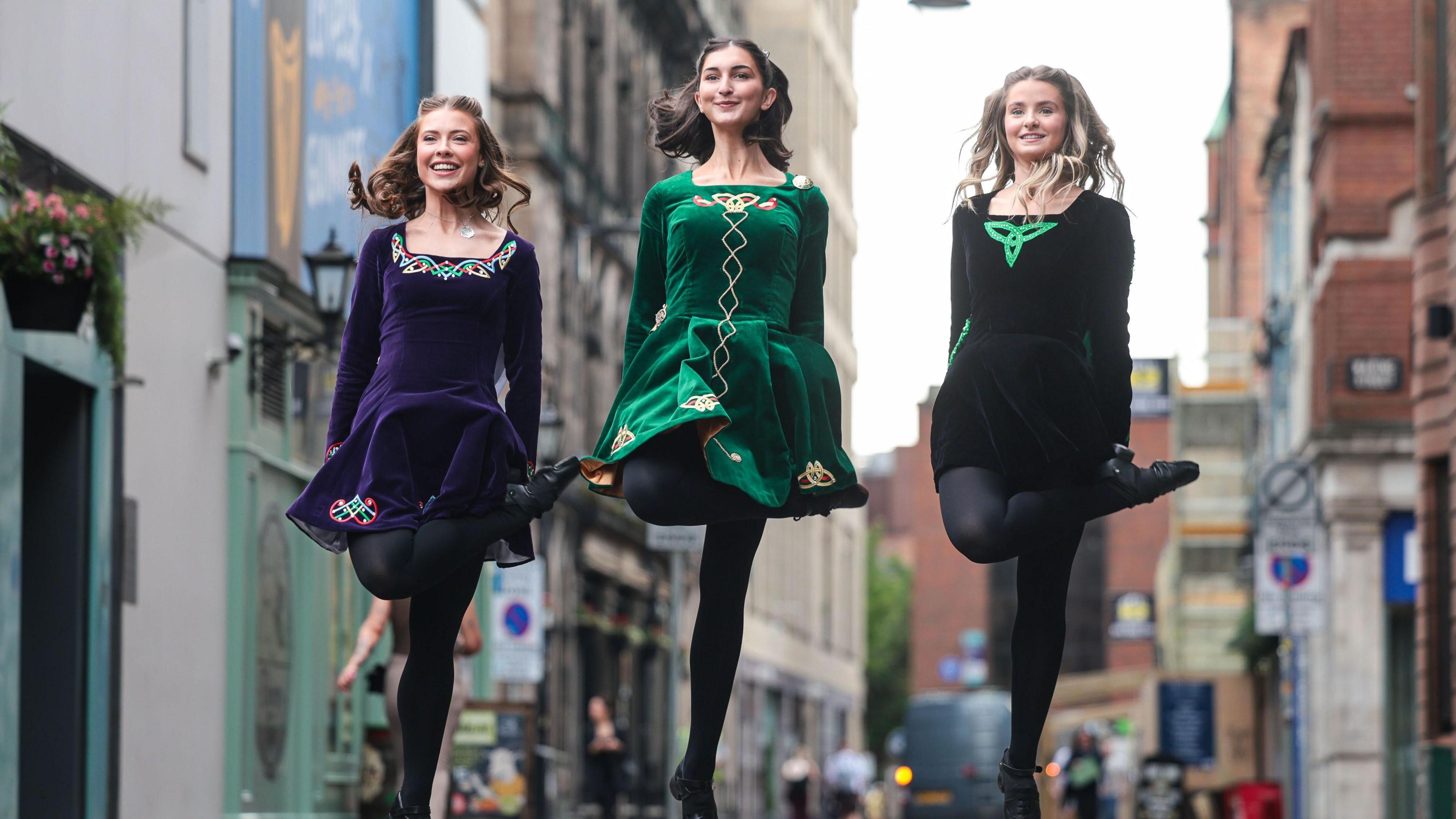 Three women in traditional Irish dancing dresses - green jewel toned velvet with Celtic embellishments. They are pictured dancing, mid leap in the air on a city street.