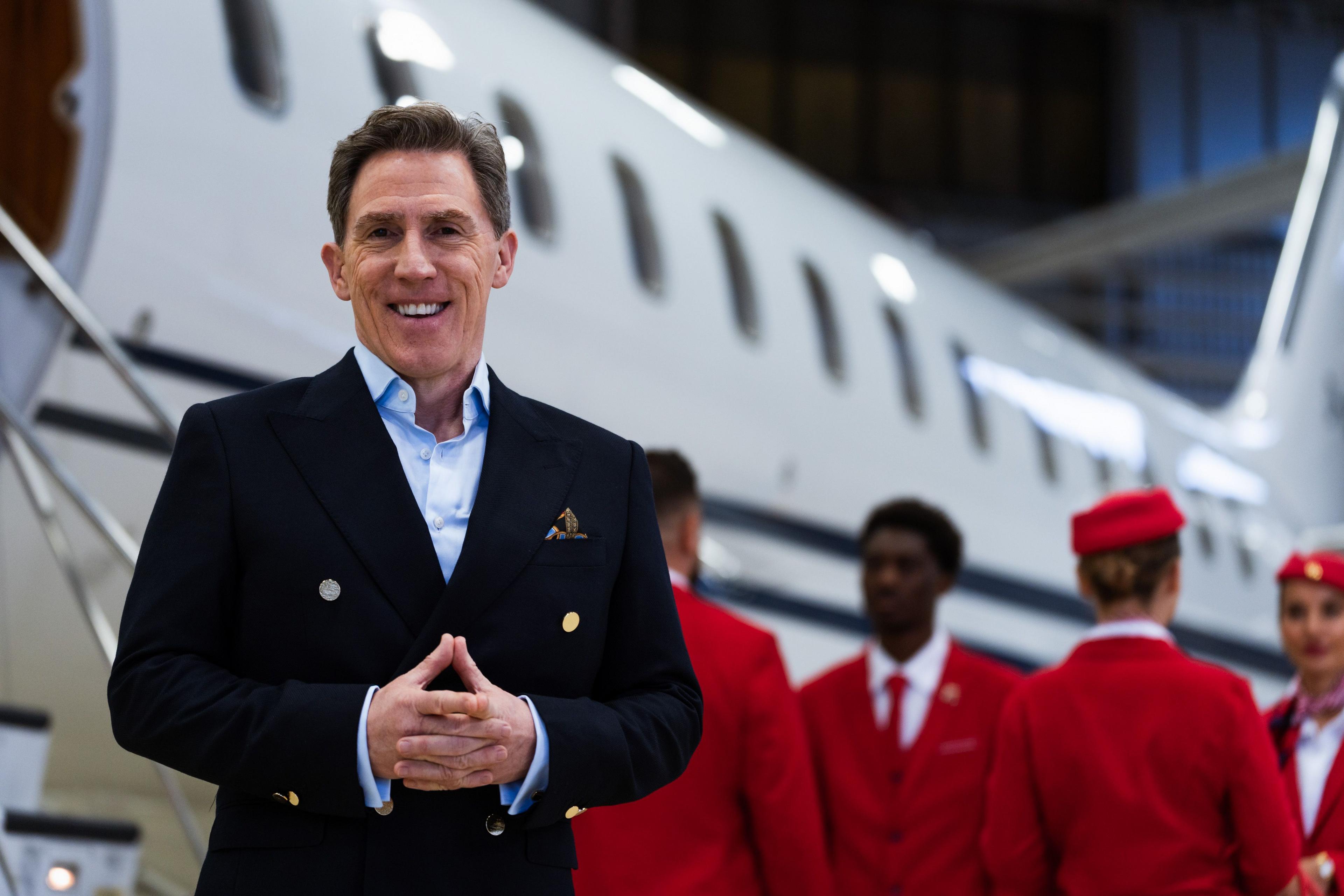 the presenter rob brydon stands in front of four members of cabin crew wearing red uniforms and a passenger jet. rob is wearing a navy suit and light blue shirt