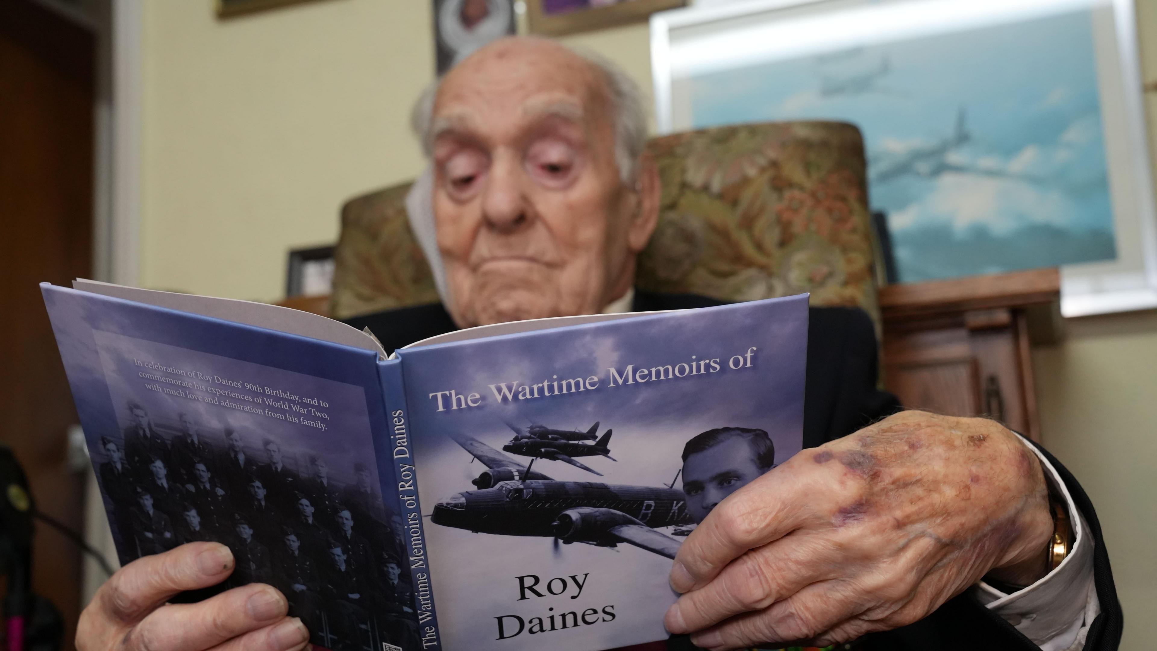 Roy Daines sat in a floral chair. He is reading a book called "The wartime memoirs of Roy Daines", with planes and his own face on the front cover.