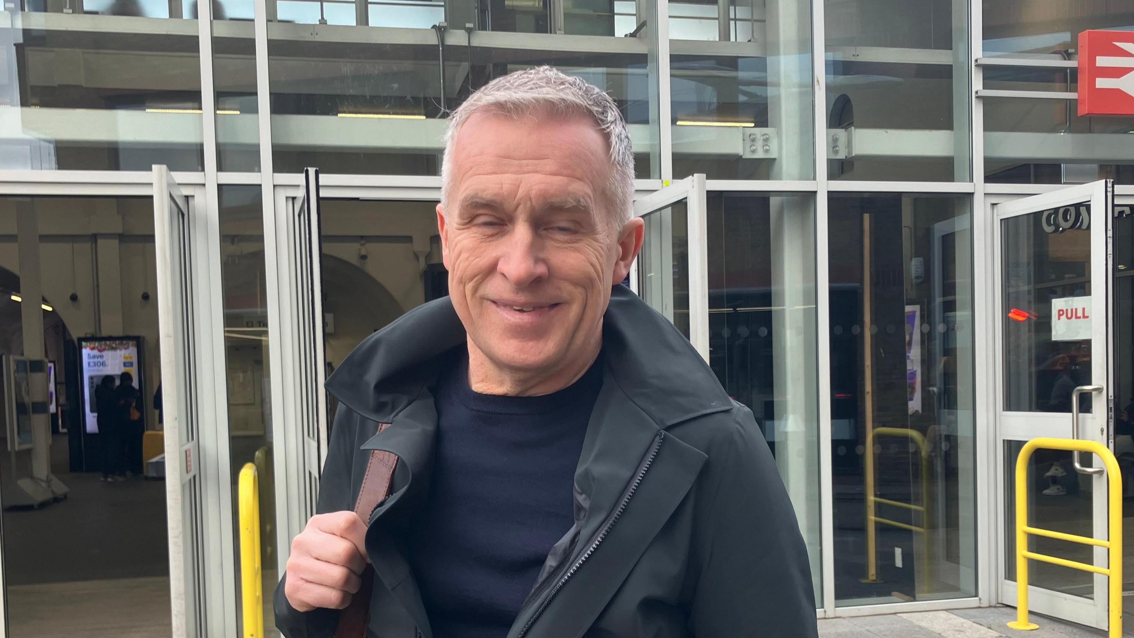 A man stands in front of Chelmsford station. He is wearing a navy jumper, black jacket and has a tan bag over one shoulder. 