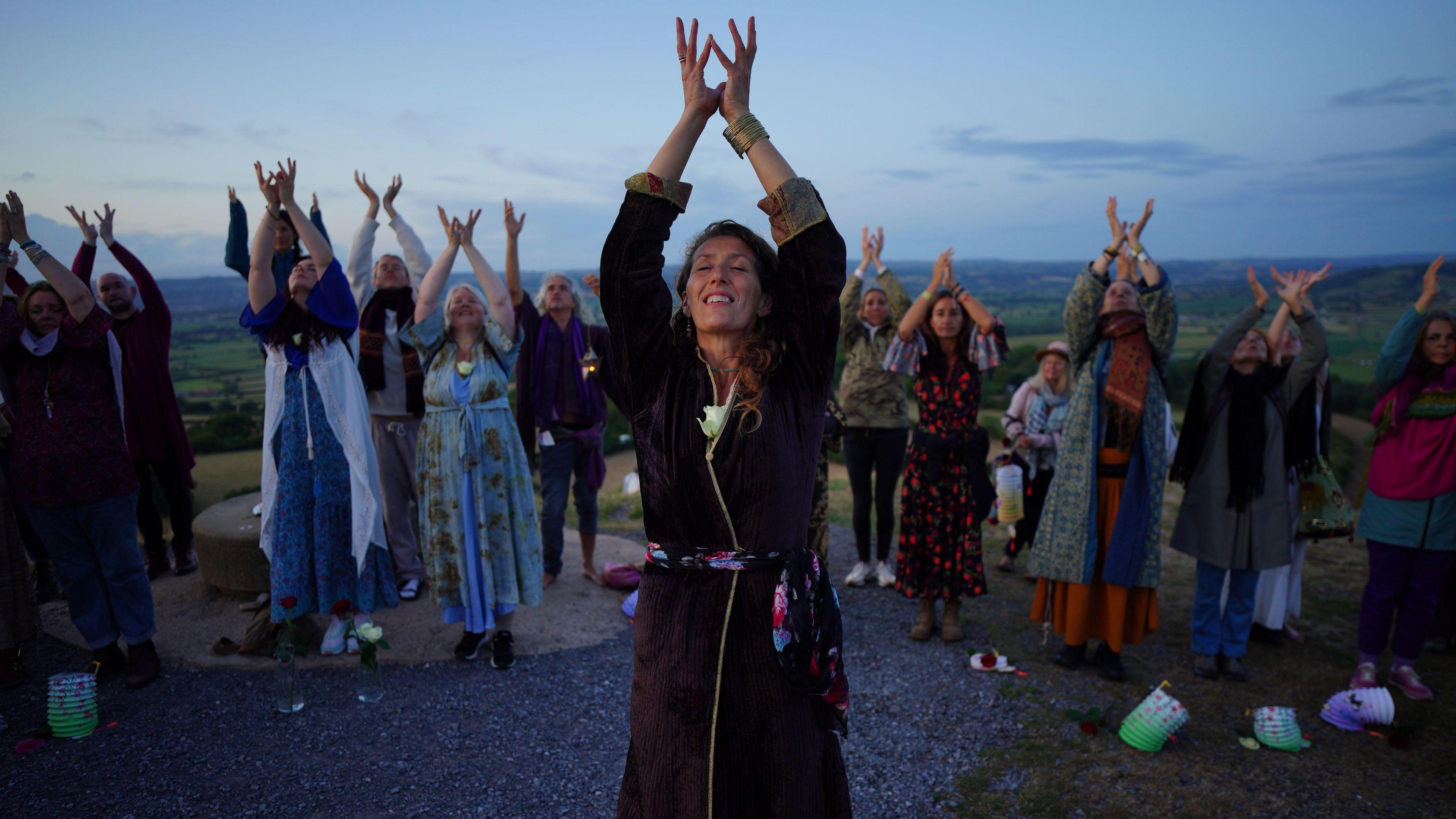 A woman in a tunic is raising her hands to the sky. Behind her is a group of people copying her movements. They are atop a hill as the sun is rising.