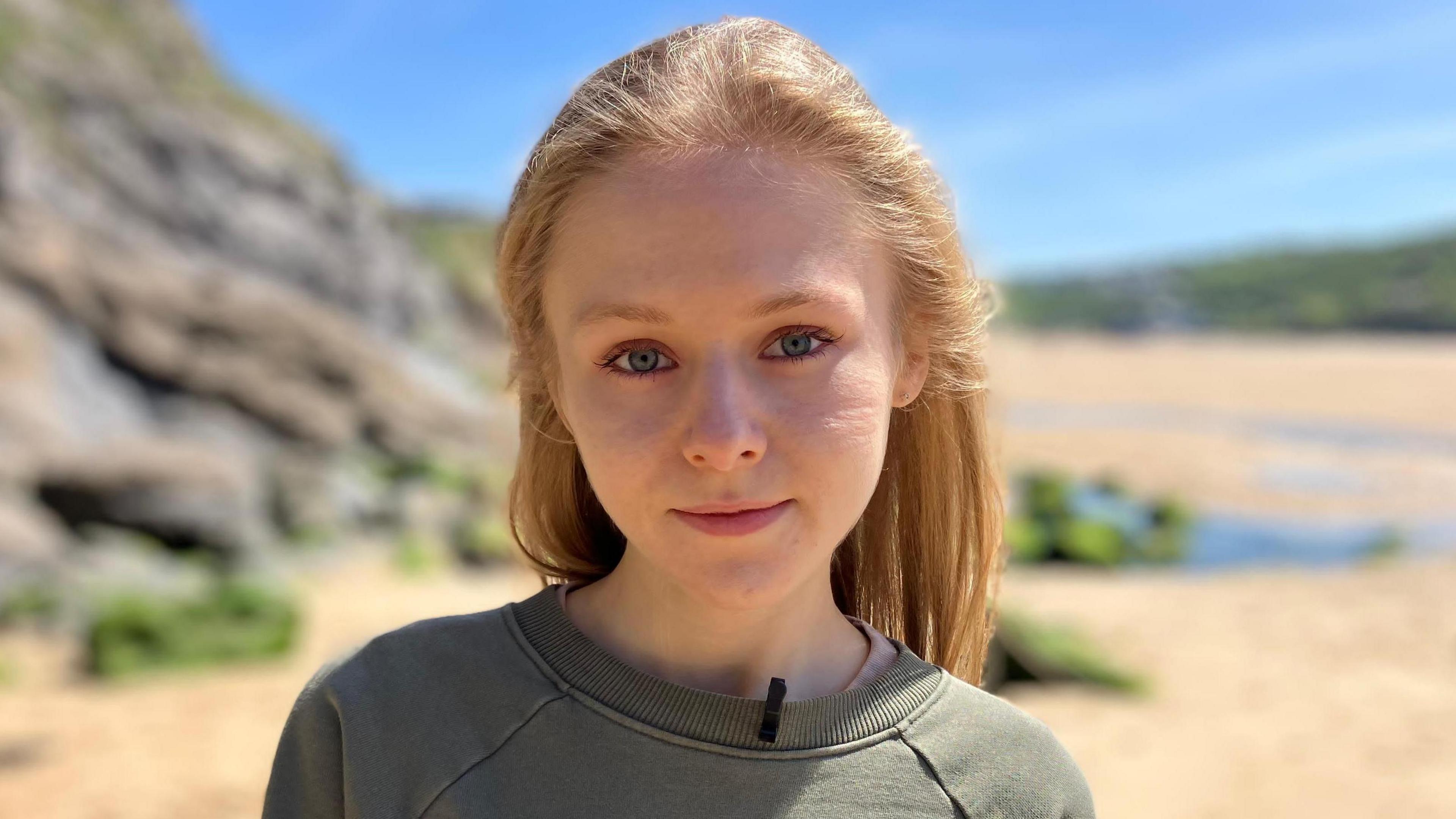 A blonde young woman with blue eyes and wearing a grey sweatshirt looks into the camera. She is standing on a beach in Cornwall and there is blue sky behind her.