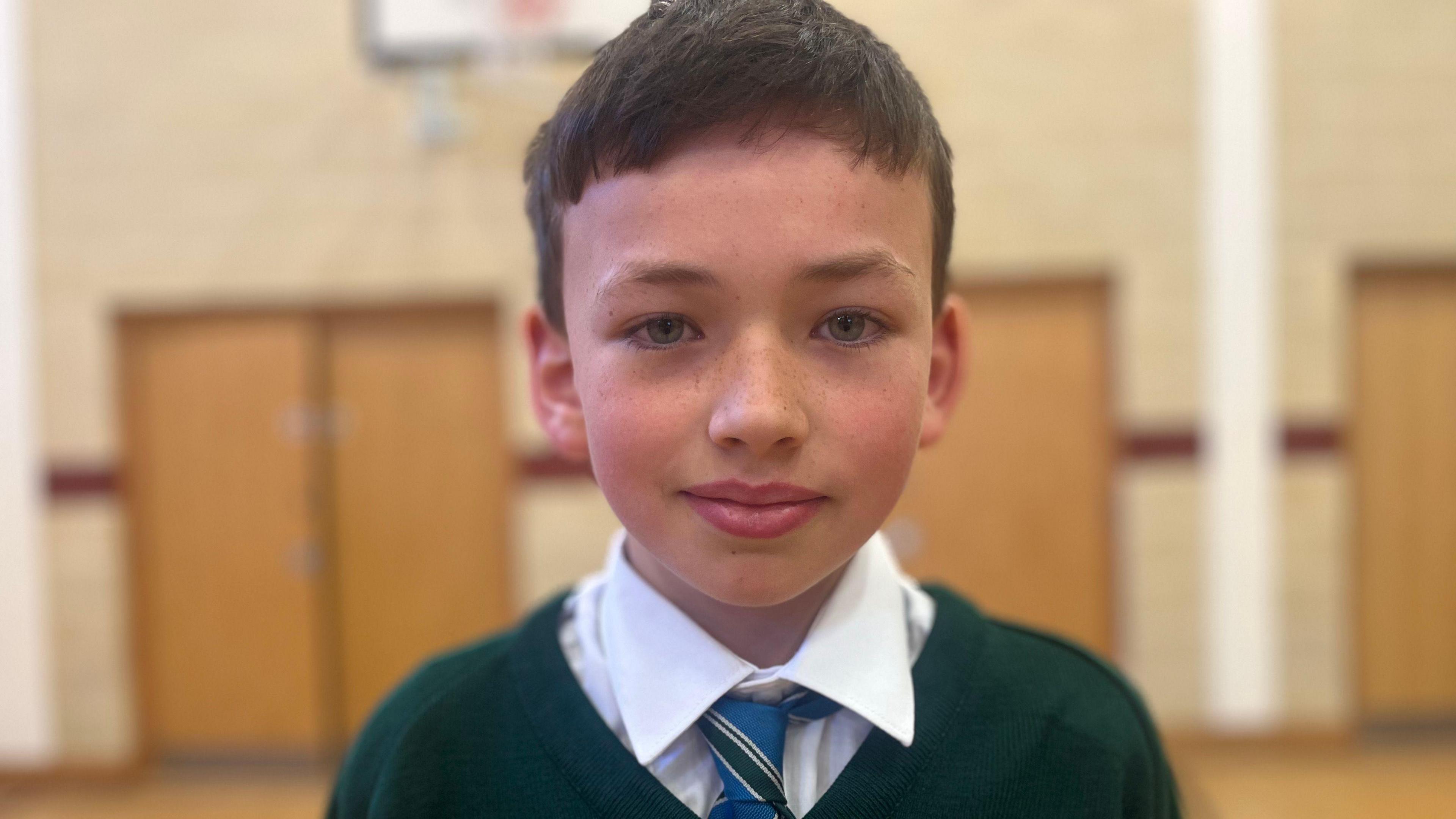 A boy is looking into the camera. He has brown hair. His uniform is green. He's standing with large doors behind him.