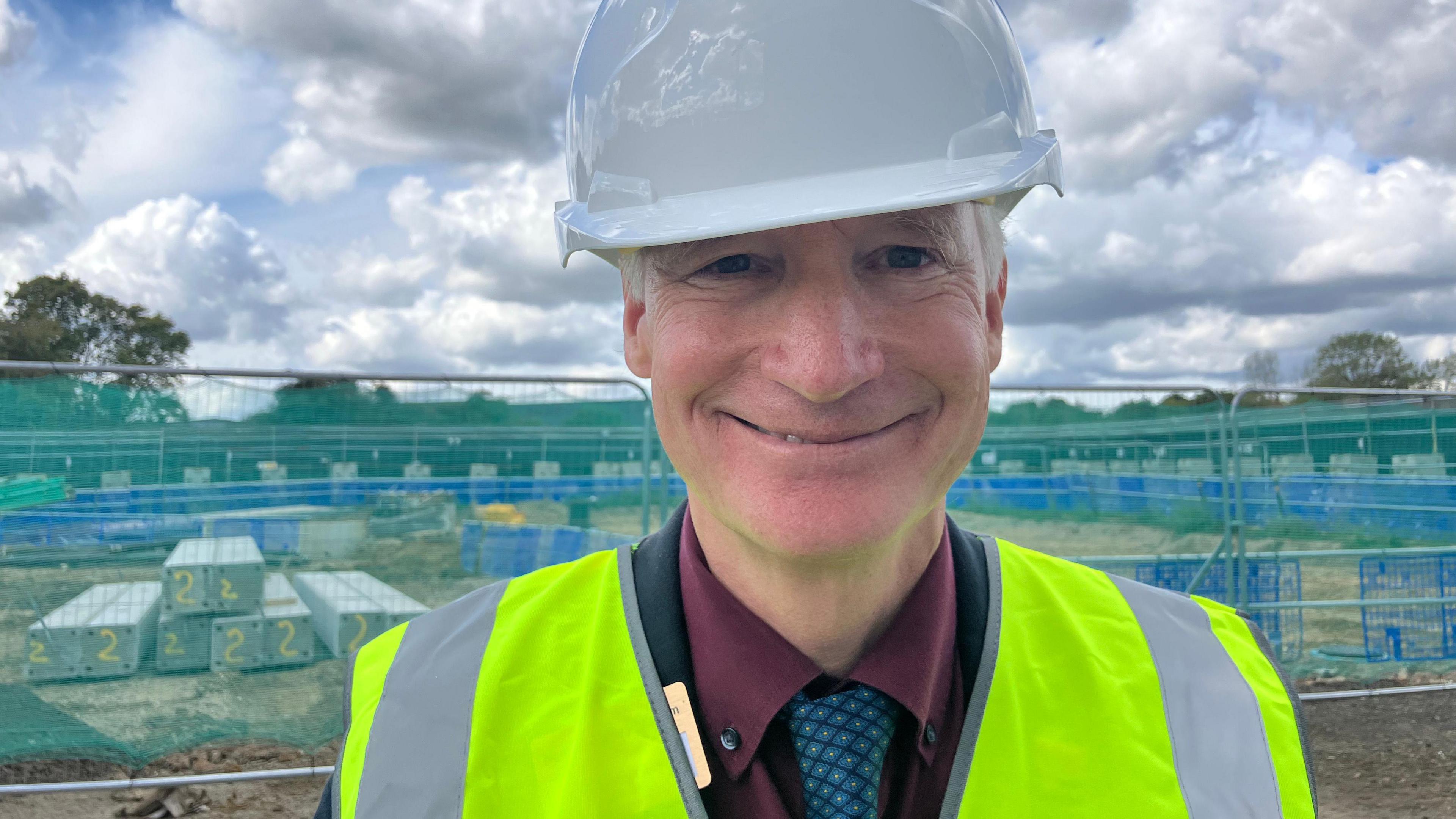 Dr Doug Gurr is wearing a high-visibility vest, a white hard had and a red shirt and blue tie. He's standing on a construction site and smiling at the camera.
